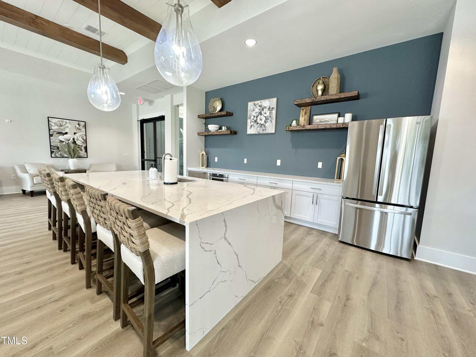 Modern kitchen with a large marble island, wicker barstools, stainless steel fridge, blue accent wall, floating shelves, and pendant lights. Light wood floors and exposed ceiling beams add warmth.