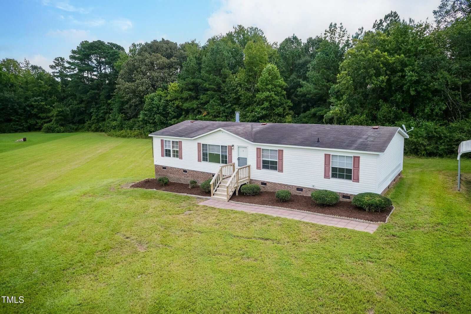Single-story white manufactured home with a dark roof, red shutters, and front steps, situated on a large grassy lot with trees in the background under a partly cloudy sky.