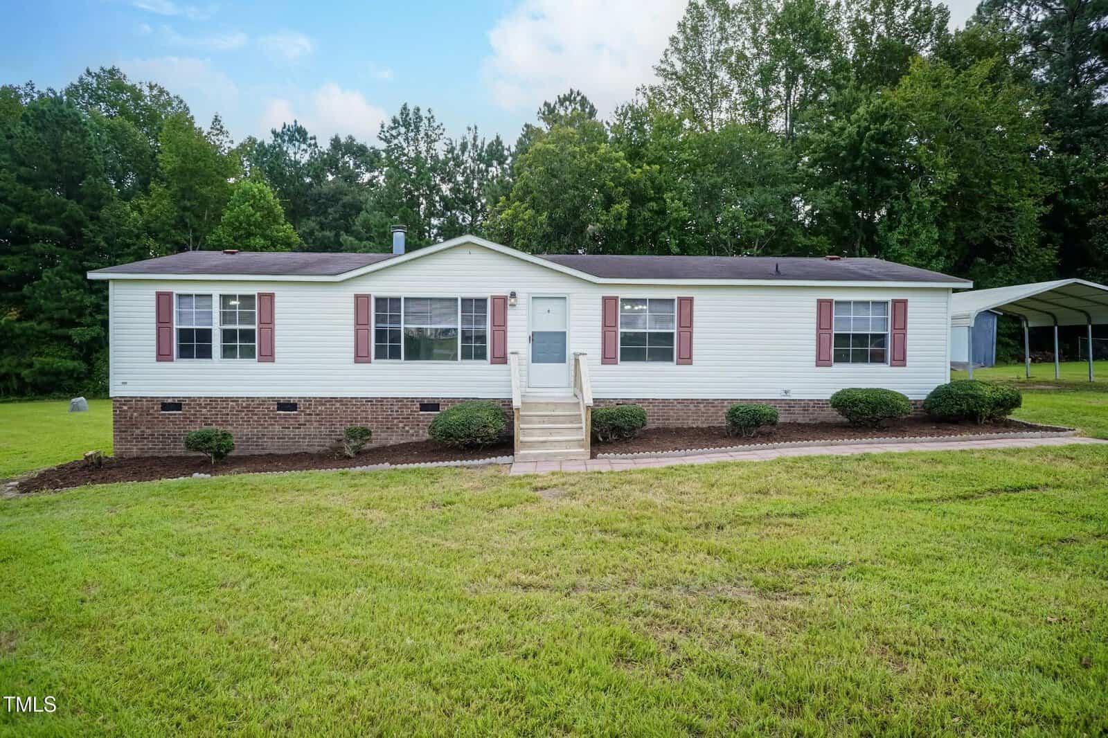 A single-story manufactured home with white siding, red shutters, and a brick foundation. There are small bushes along the front, concrete steps with railings, and a grassy yard with trees in the background.