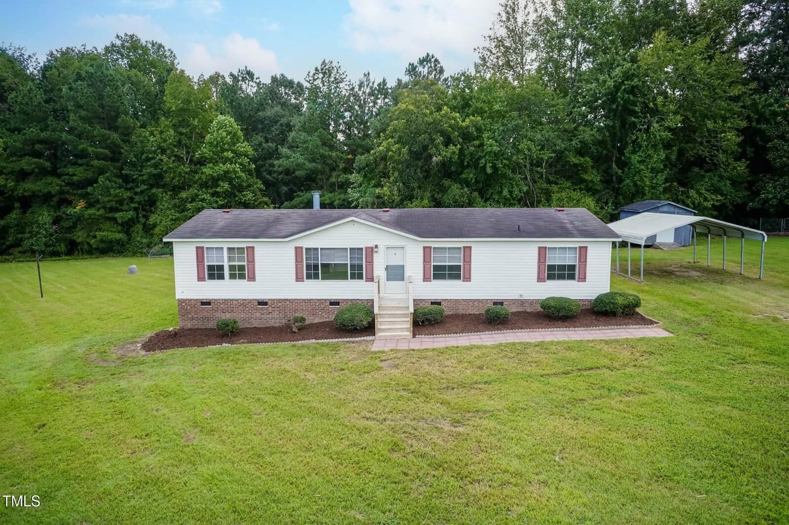 Charming single-story white manufactured home with red shutters, surrounded by green grass and bordered by bushes in front. Located at 40 Beaver Run in Spring Hope, this property features dense trees behind the home, and a metal carport is seen at the back right of the property.