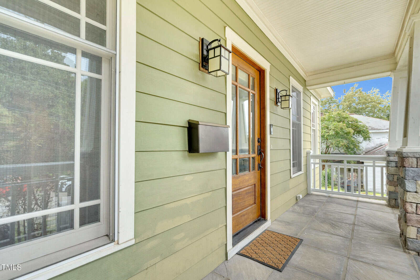 A green house front porch with tile flooring, a wooden door with glass panes, two black wall lanterns, a black mailbox, and a welcome mat. A white-trimmed window and part of a stone column are also visible.