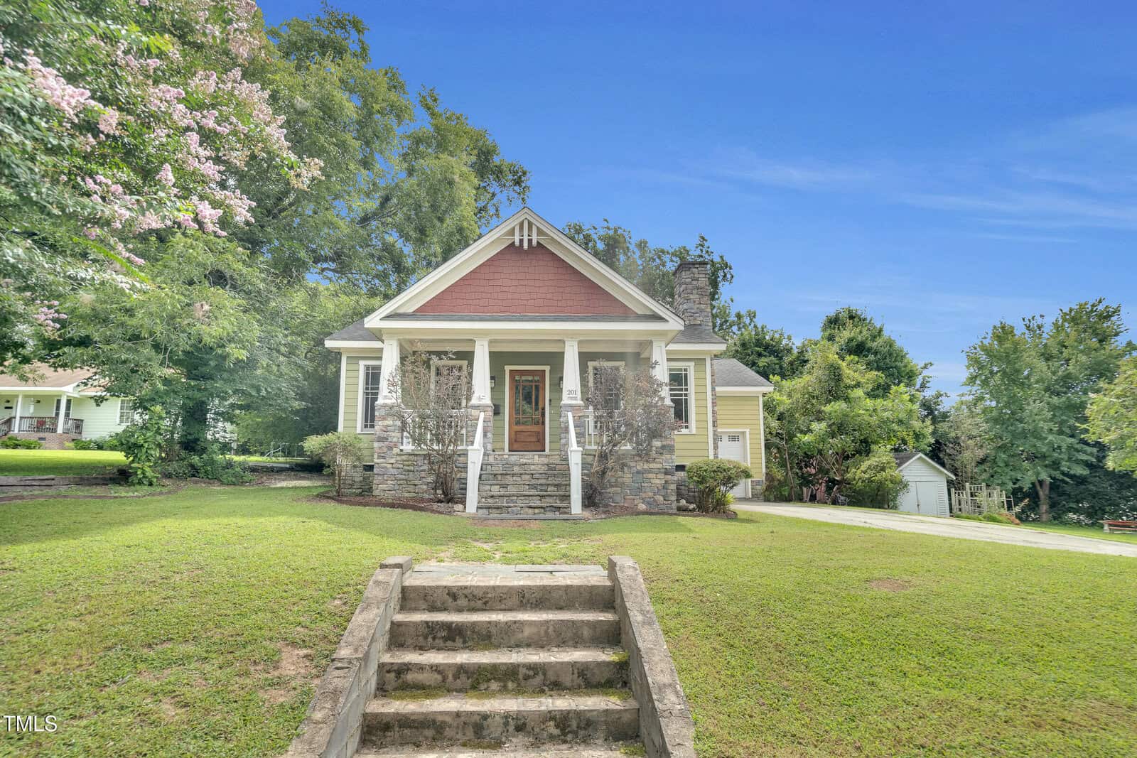 A small house with a red gable roof, stone steps leading to a covered porch with columns, and a well-maintained lawn. Trees surround the property at 201 Cedar Street, Louisburg, and a driveway is visible on the right side.