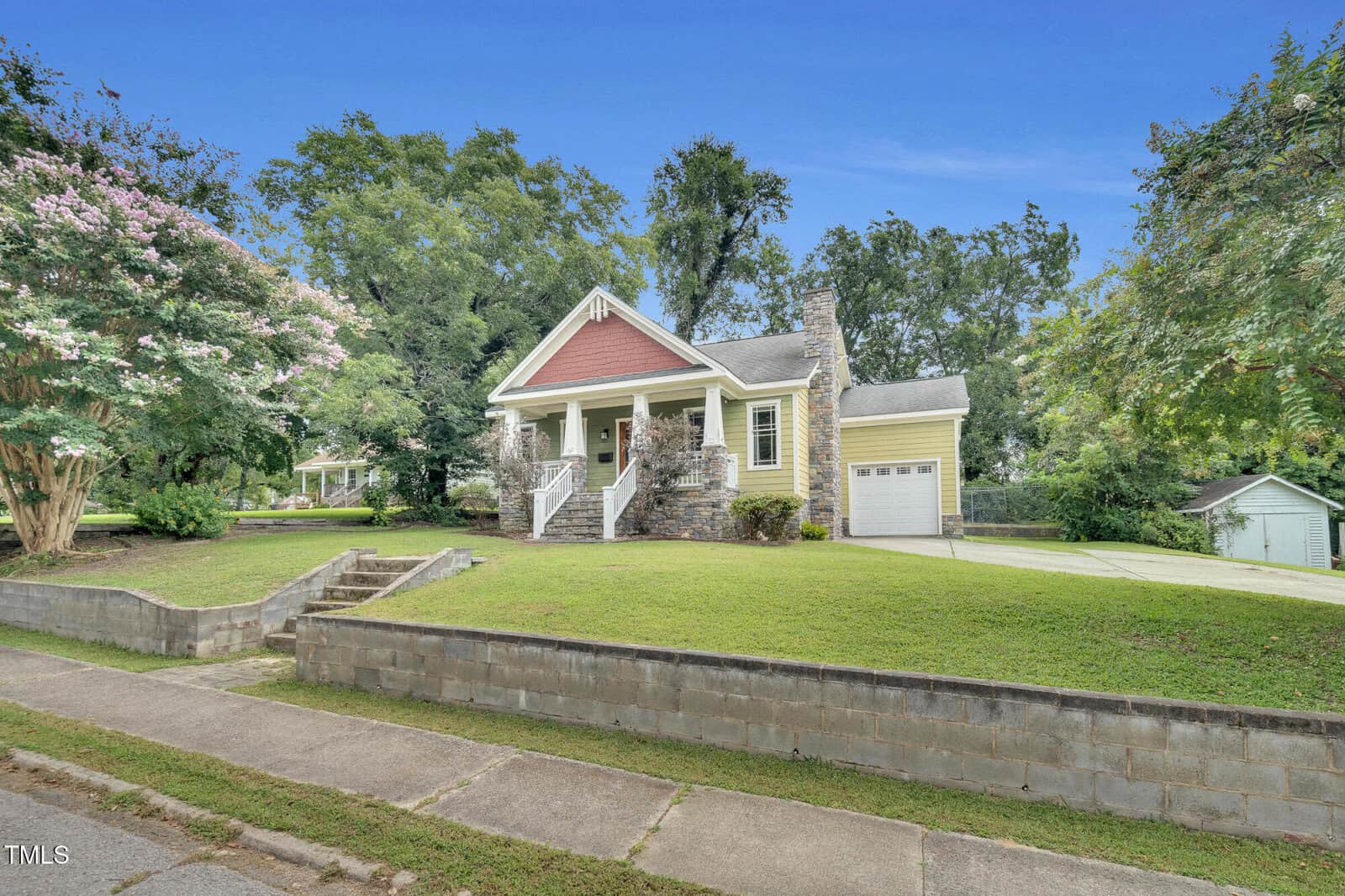 A yellow house with a red gabled porch, stone accents, white railings, and an attached garage sits on a grassy lawn with mature trees and a concrete driveway, viewed from the street on a clear day.