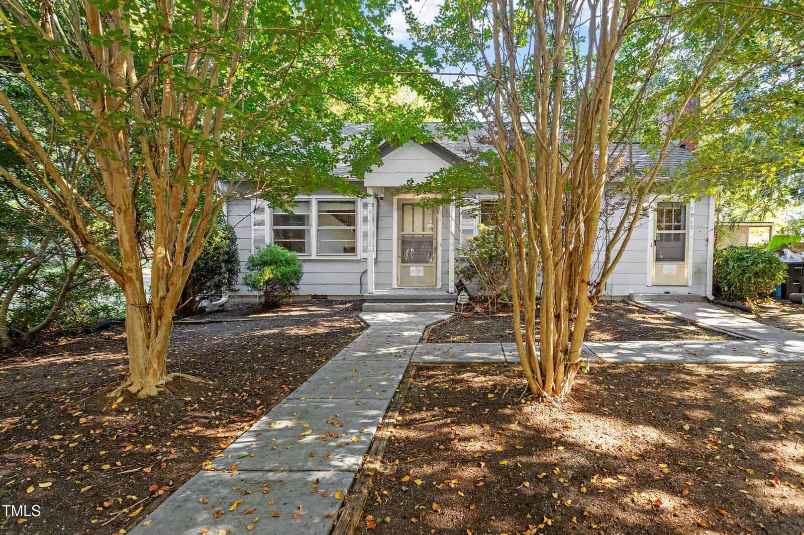 A small, white, single-story house with a gray roof at 5063 Nc-56 Hwy, Franklinton is partially obscured by trees. A concrete walkway leads from the sidewalk to the front door. The yard features mulched soil and sparse fallen leaves.