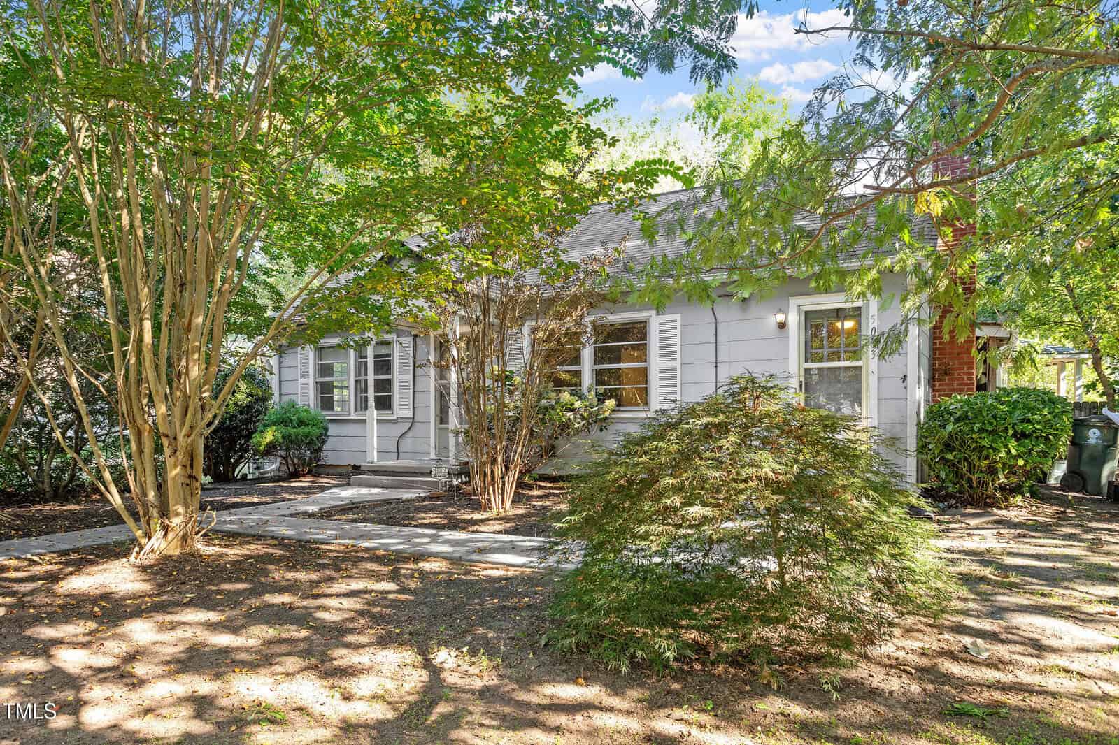 A small gray house with white shutters is partially shaded by trees and surrounded by foliage. A sidewalk leads to the front door, and sunlight filters through the leaves, casting shadows on the ground.