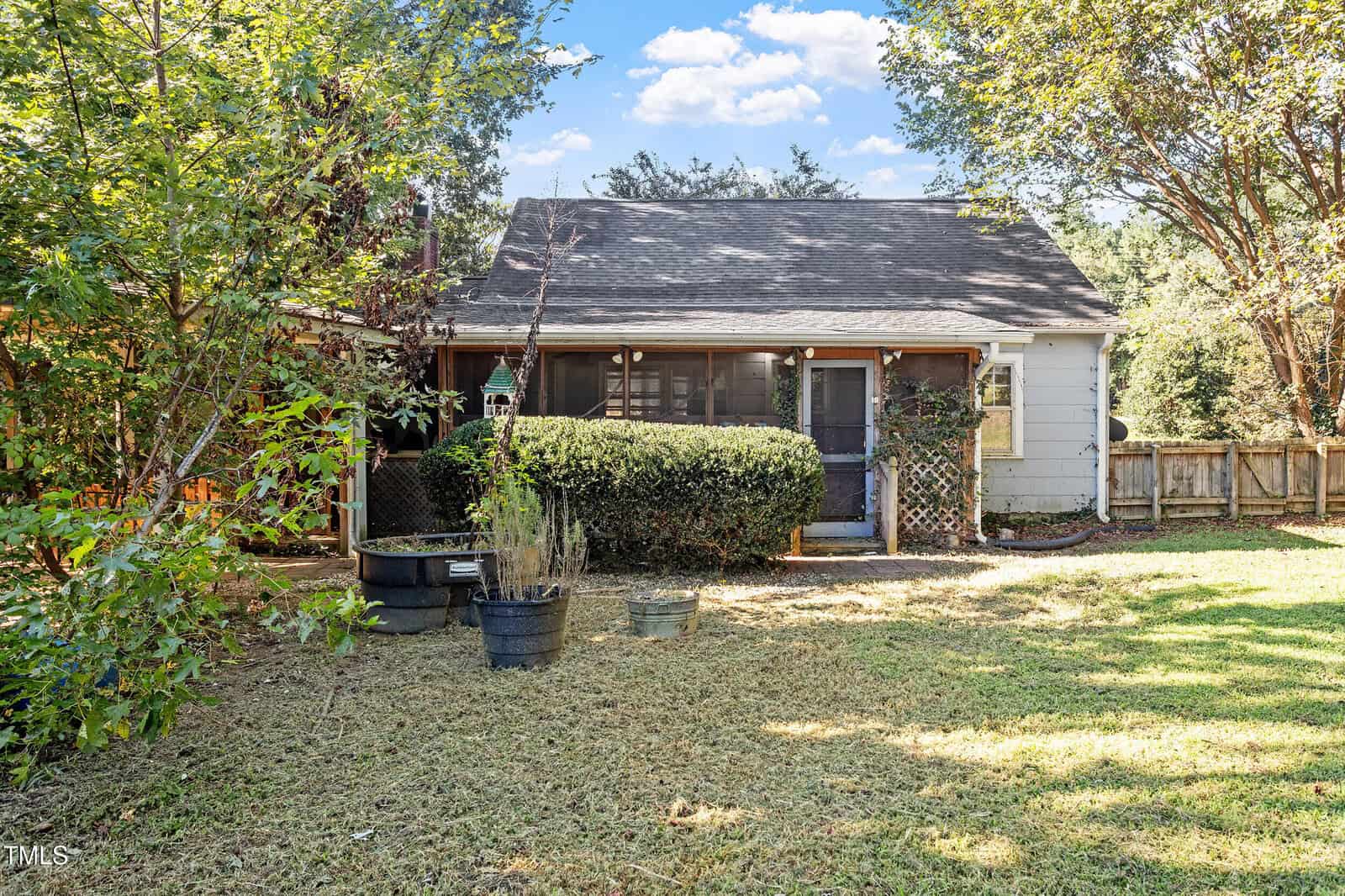 A small house with a shingled roof, a screened-in porch, and a hedge in front. Potted plants sit on the lawn, and a wooden fence and trees surround the grassy yard under a blue sky.