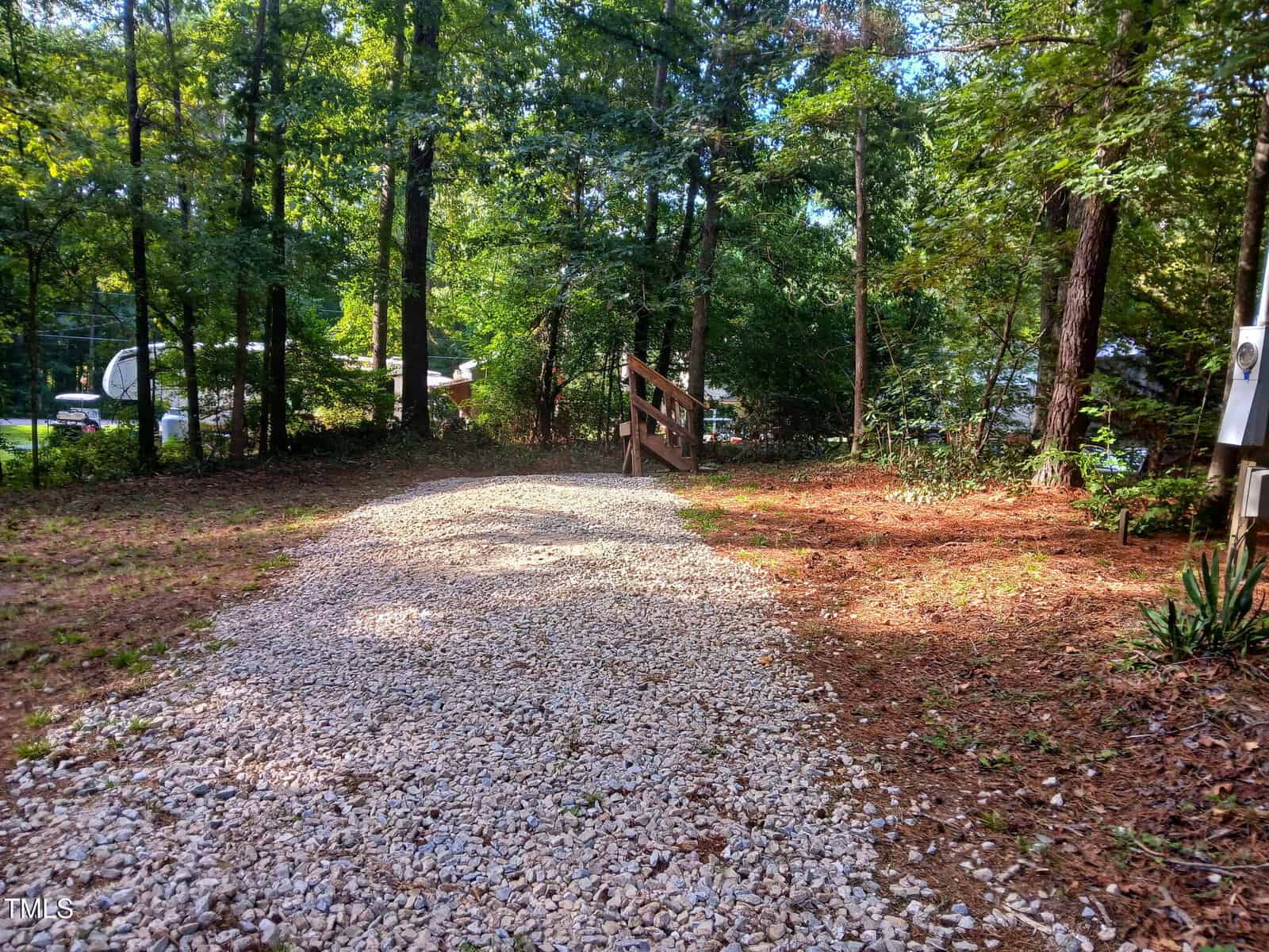 A gravel driveway curves through a wooded area with tall trees on both sides. Sunlight filters through the leaves, and a wooden handrail borders a small incline in the background.