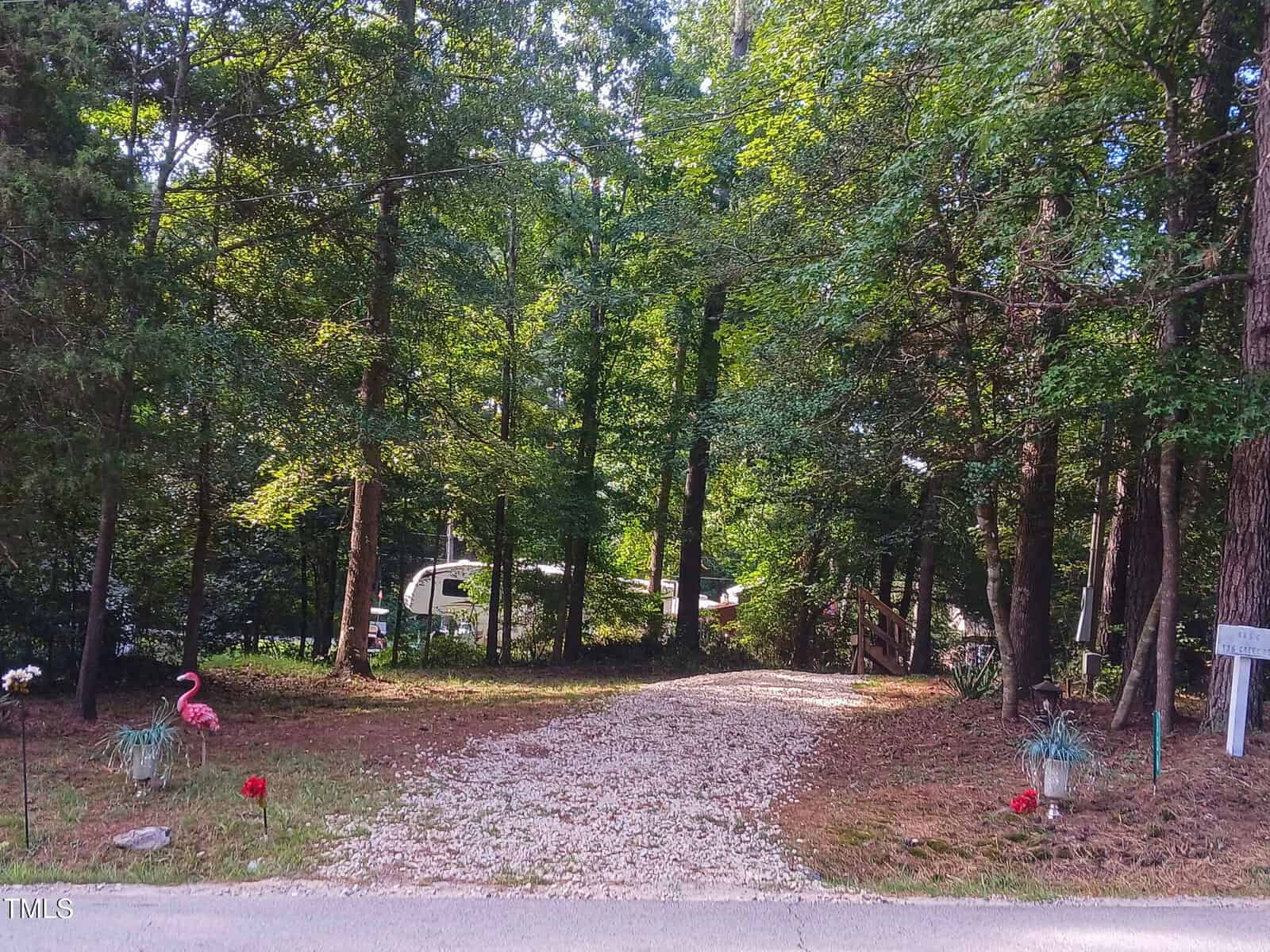 A gravel driveway leads through a wooded area toward a white RV, with potted plants, a pink flamingo lawn ornament, and mailboxes visible near the entrance. Sunlight filters through the dense green trees.