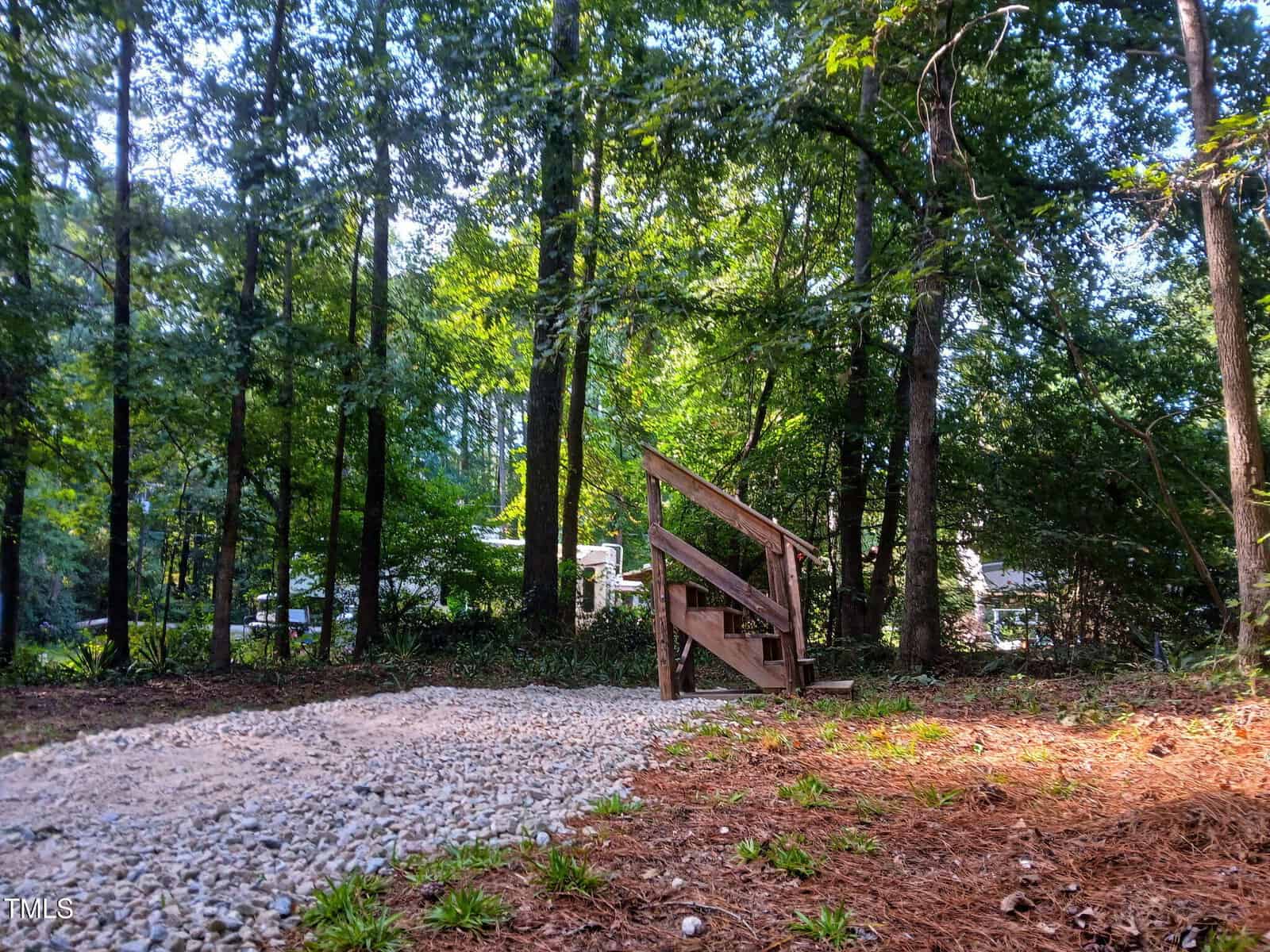 A small wooden staircase leads from a gravel path into a wooded area with tall trees and dense greenery. Sunlight filters through the leaves, and a building is visible in the background.