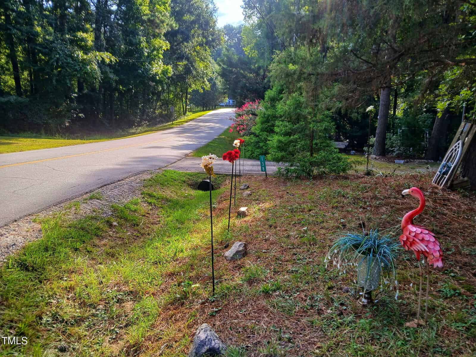 A quiet rural roadside with tall trees, a grassy ditch, artificial flowers on stakes, a pink plastic flamingo, and a potted plant, all bathed in bright morning sunlight.