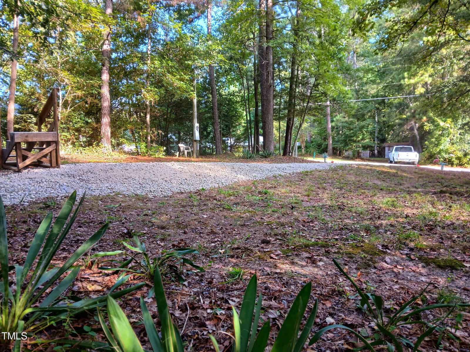 A gravel driveway curves through a wooded area with tall trees and scattered sunlight. A white car is parked near the road, and some wooden steps and greenery are visible in the foreground.