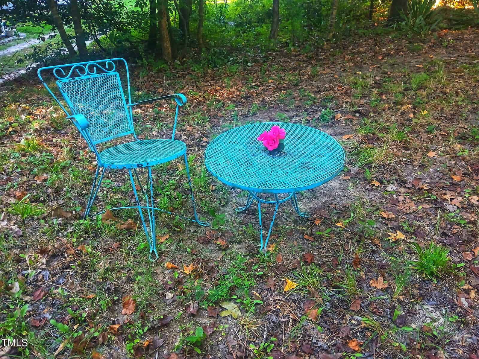 A blue metal chair and round table sit on a patch of grass and dirt in a wooded area; a small pink flower pot is placed on the table.