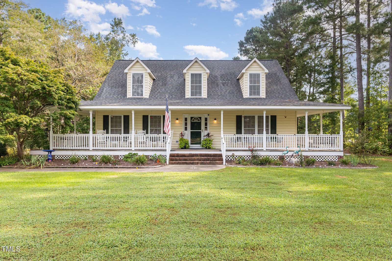 A charming, two-story house at 30 Beddingfield Drive, Zebulon, with three dormer windows, a covered wraparound porch, white railings, and a central front door surrounded by a green lawn, trees, and landscaped flowerbeds.