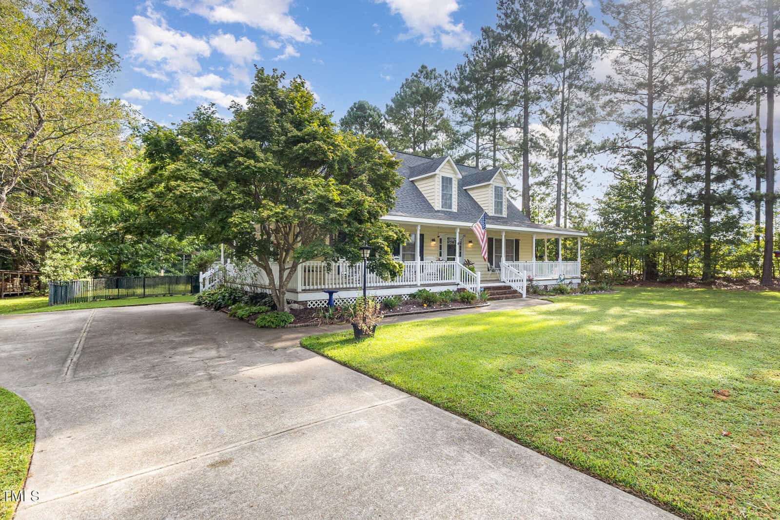 A large house with a wraparound porch, American flag, and a sloped driveway. The home is surrounded by green grass, trees, and landscaped plants under a partly cloudy sky.