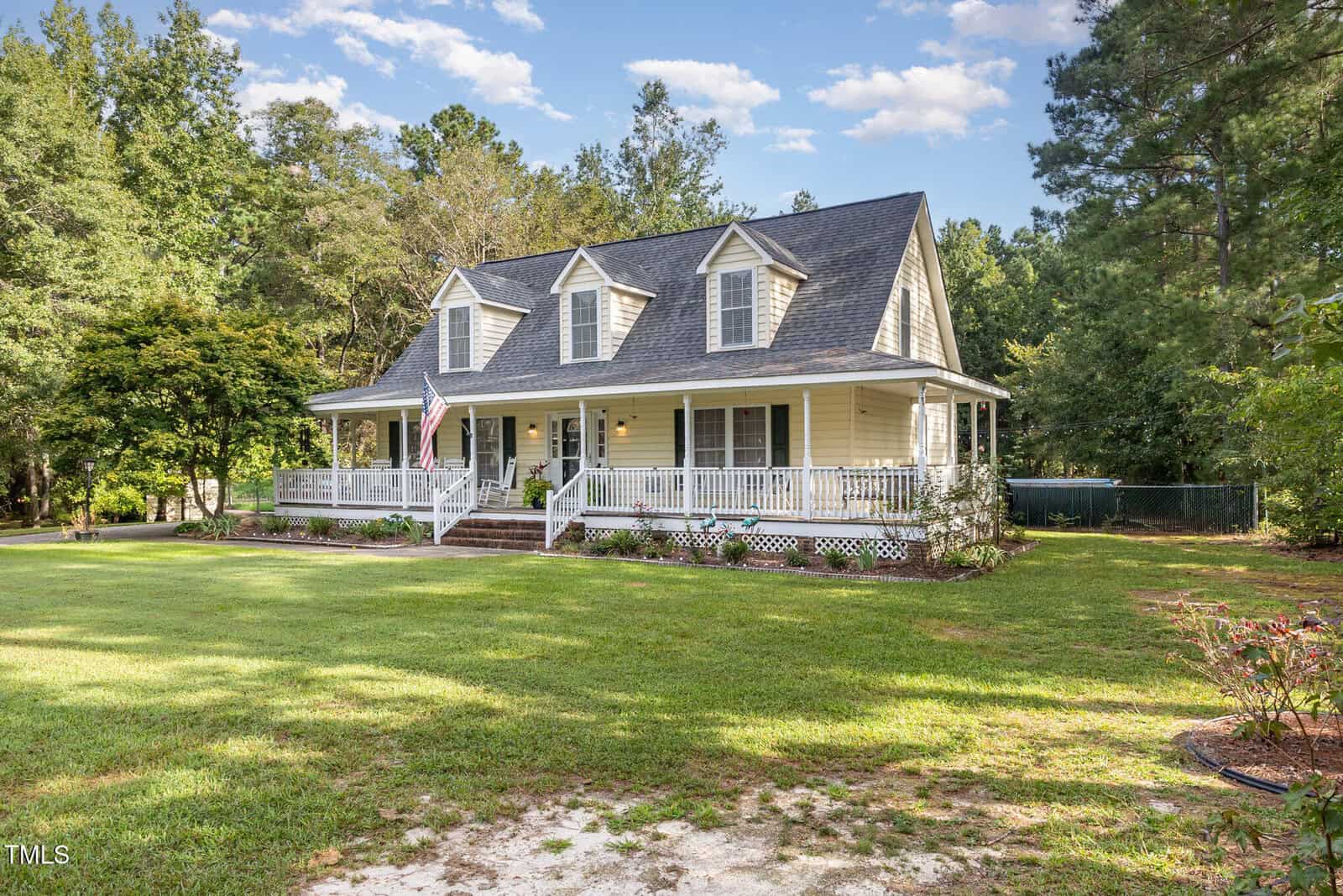 A charming yellow house with a large front porch, white railings, and dormer windows, surrounded by grass, trees, and a flag by the steps under a blue sky with scattered clouds.