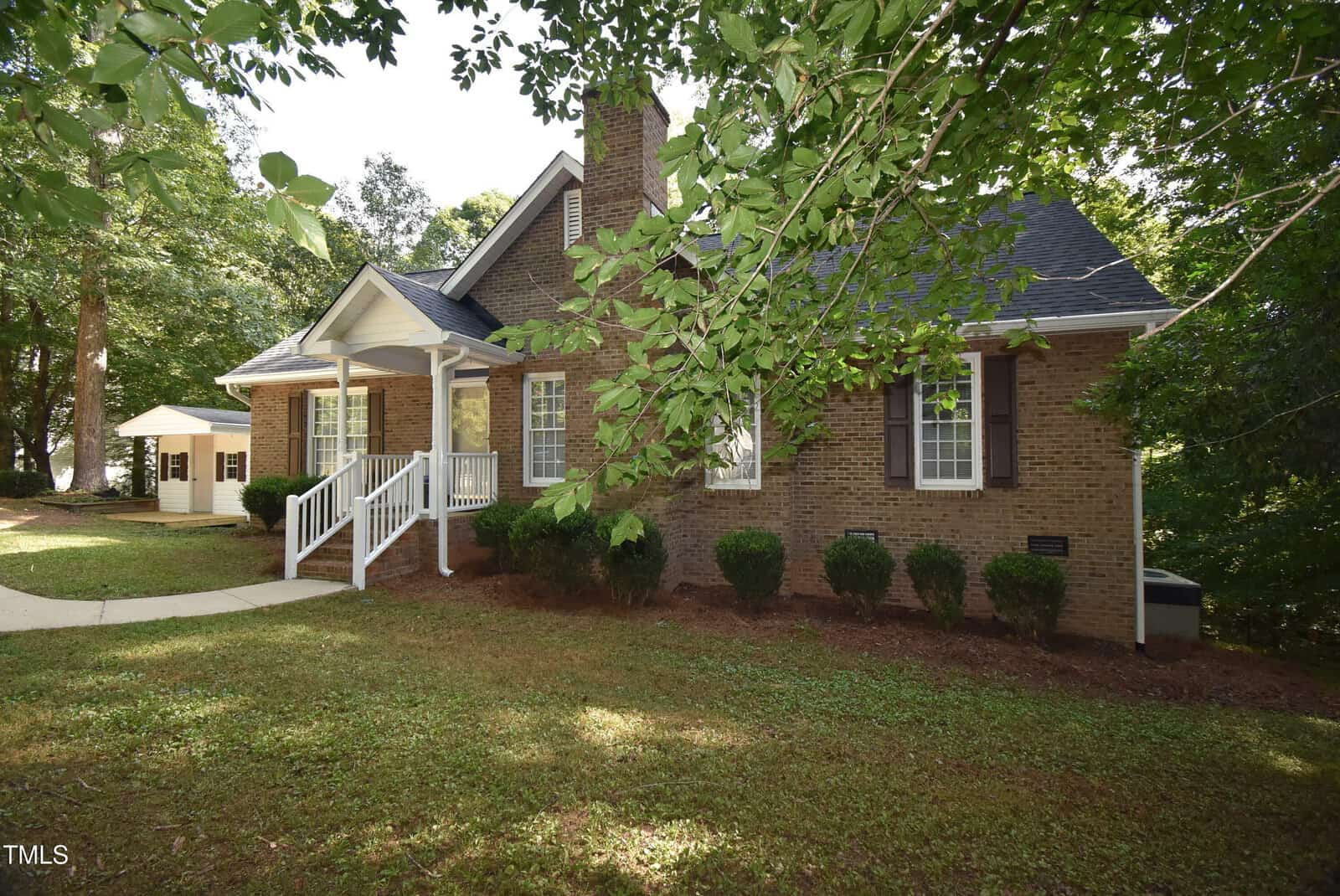 A brick house at 205 Fox Run Road in Youngsville features a porch with white railing, dark shutters, and is surrounded by trees and bushes, with a small white shed in the background and a curved sidewalk leading to the entrance.