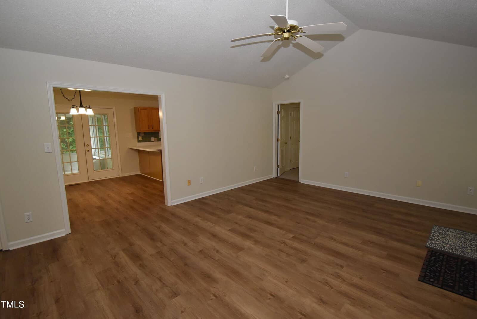 A spacious living room with wood flooring, cream-colored walls, a vaulted ceiling with a ceiling fan, and an open view into a kitchen and dining area through a wide doorway.
