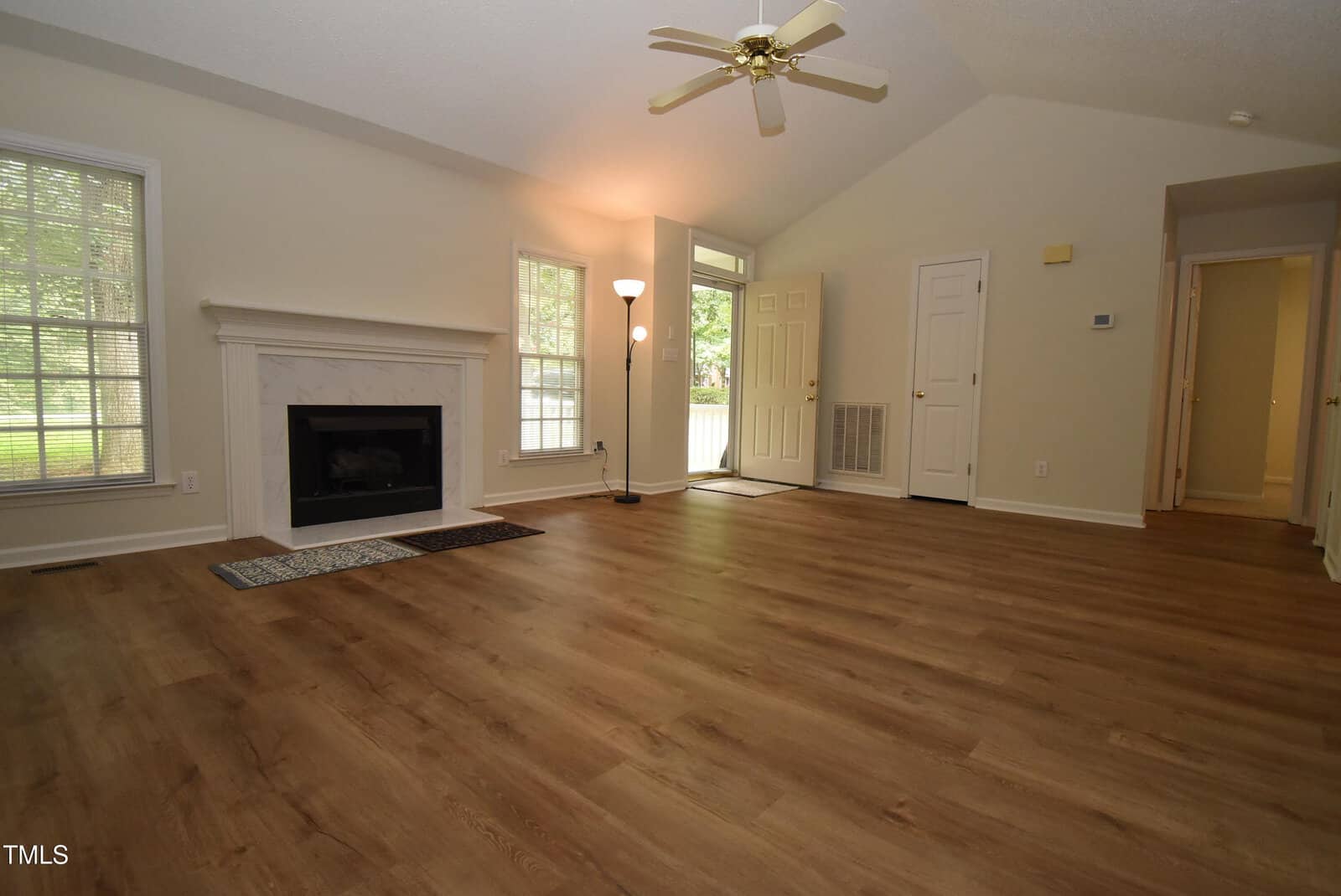 Spacious living room with wood floors, a white fireplace, large windows, a ceiling fan, and an open doorway leading to a hallway and front door. Natural light fills the room.