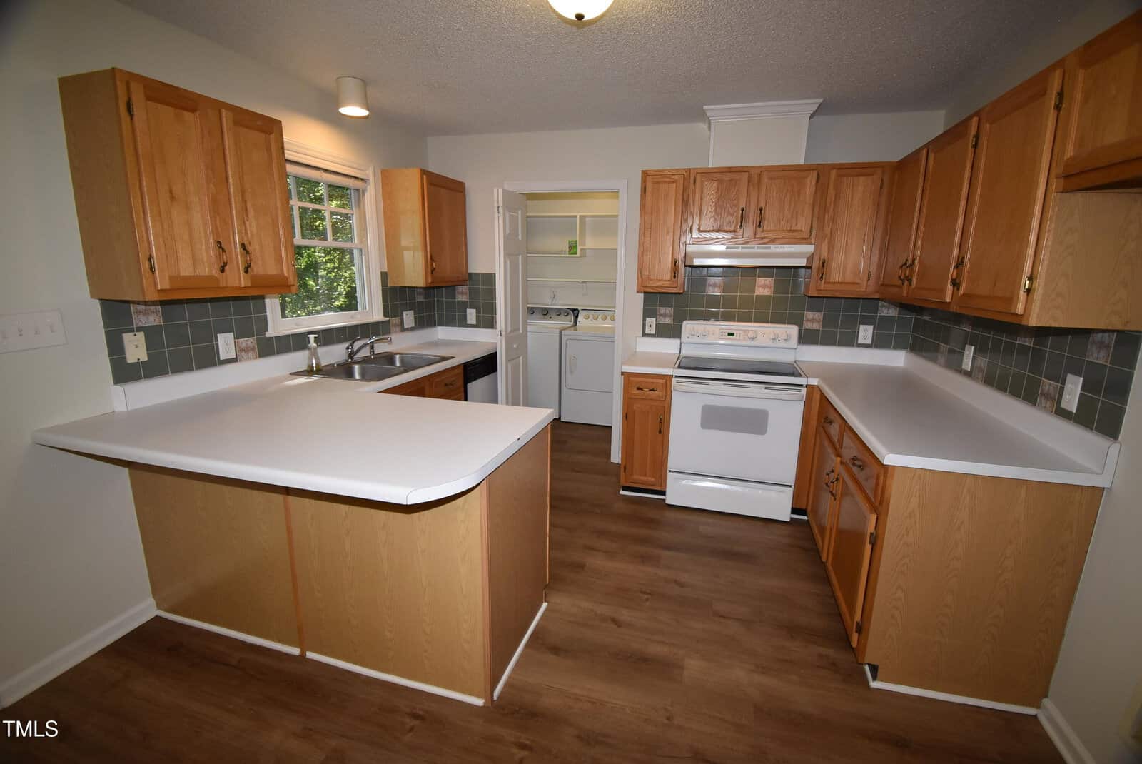 A kitchen with light wood cabinets, white countertops, and a white stove. A window above the sink lets in natural light. A laundry area with a washer and dryer is visible through an open doorway. The floor is light wood.