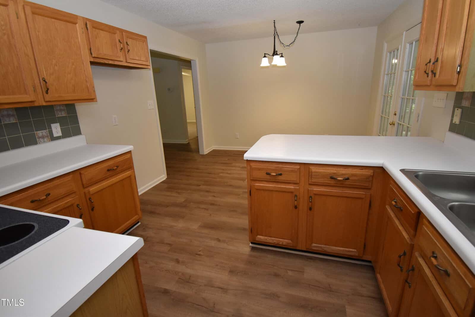 A kitchen with wooden cabinets, white countertops, a sink, stove, and green tile backsplash, opening into a dining area with wood flooring and a hanging light fixture. Double doors are visible on the right.