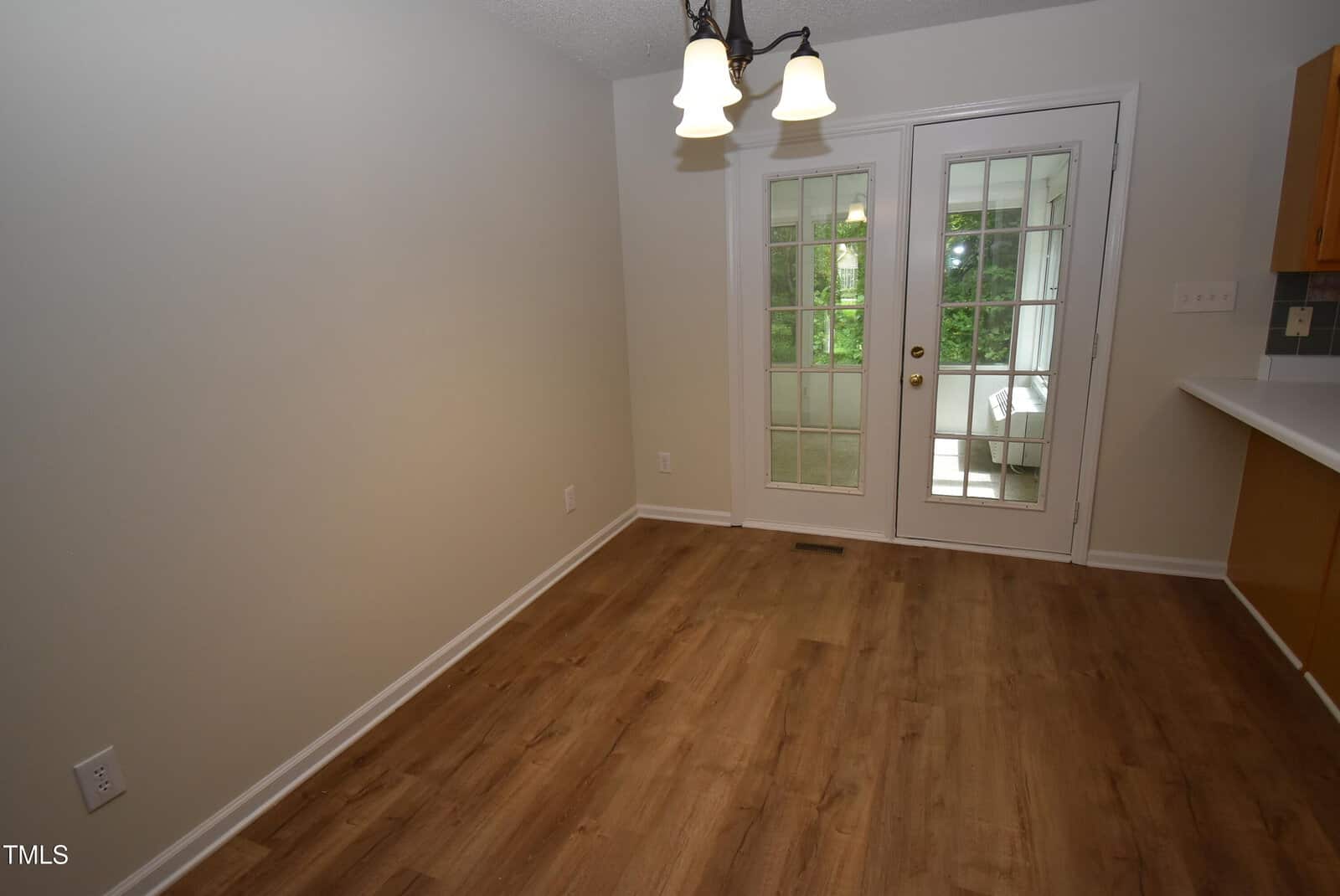 A small dining area with light wood flooring, beige walls, a white ceiling, and a hanging light fixture. French doors with glass panes lead to a bright sunroom with trees visible outside.