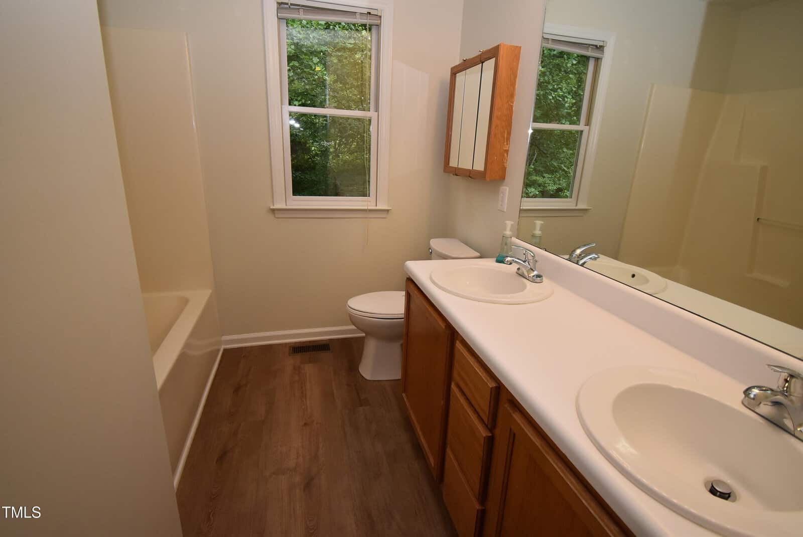 A bathroom with wood flooring, double sinks with a large mirror, a toilet next to two windows, a wooden medicine cabinet, and a bathtub with a shower in the corner.