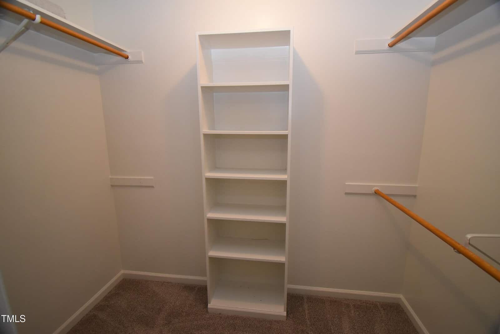 Walk-in closet with beige carpet, white walls, wooden hanging rods on both sides, and a white shelving unit in the center.