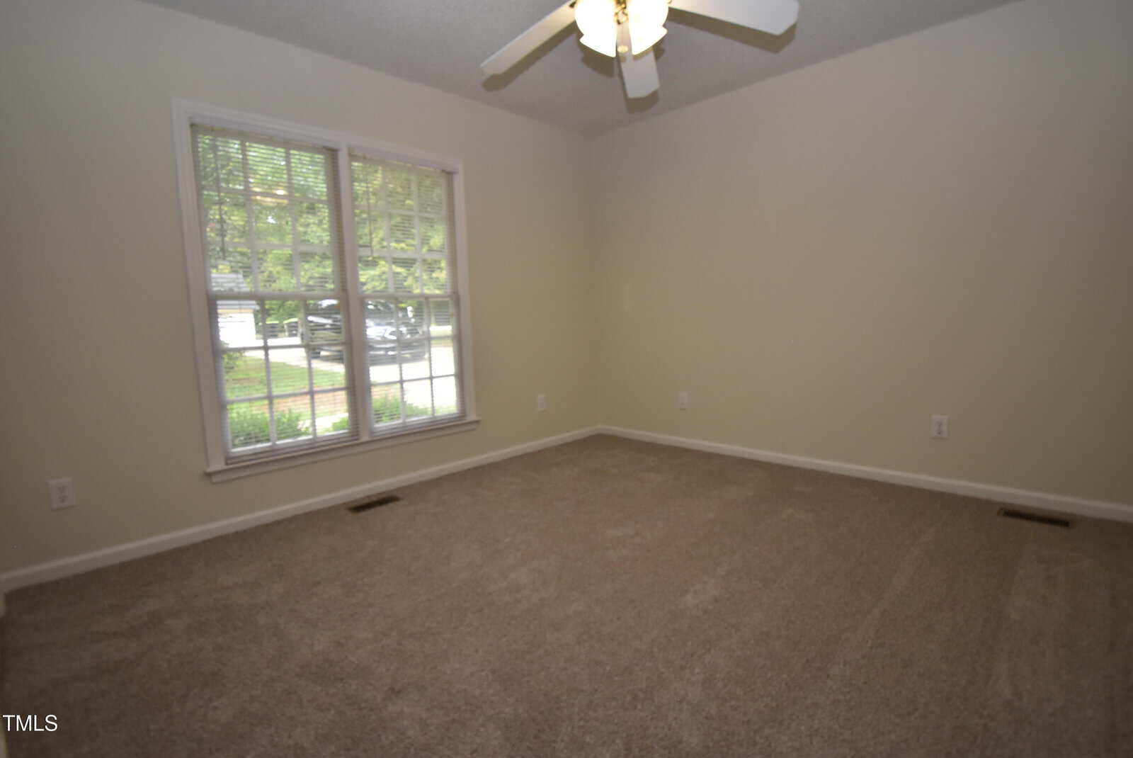 Empty carpeted room with beige walls, a large window letting in natural light, and a ceiling fan with a light fixture.