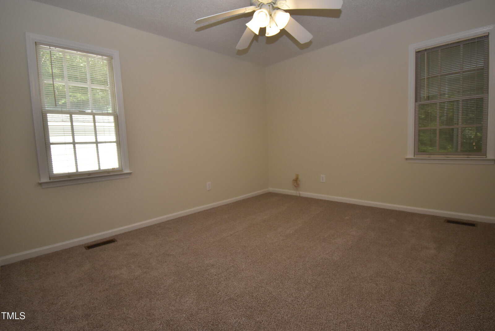 Empty carpeted room with beige walls, two windows with blinds, a ceiling fan with lights, and electrical outlets on the walls. Natural light enters through the windows.