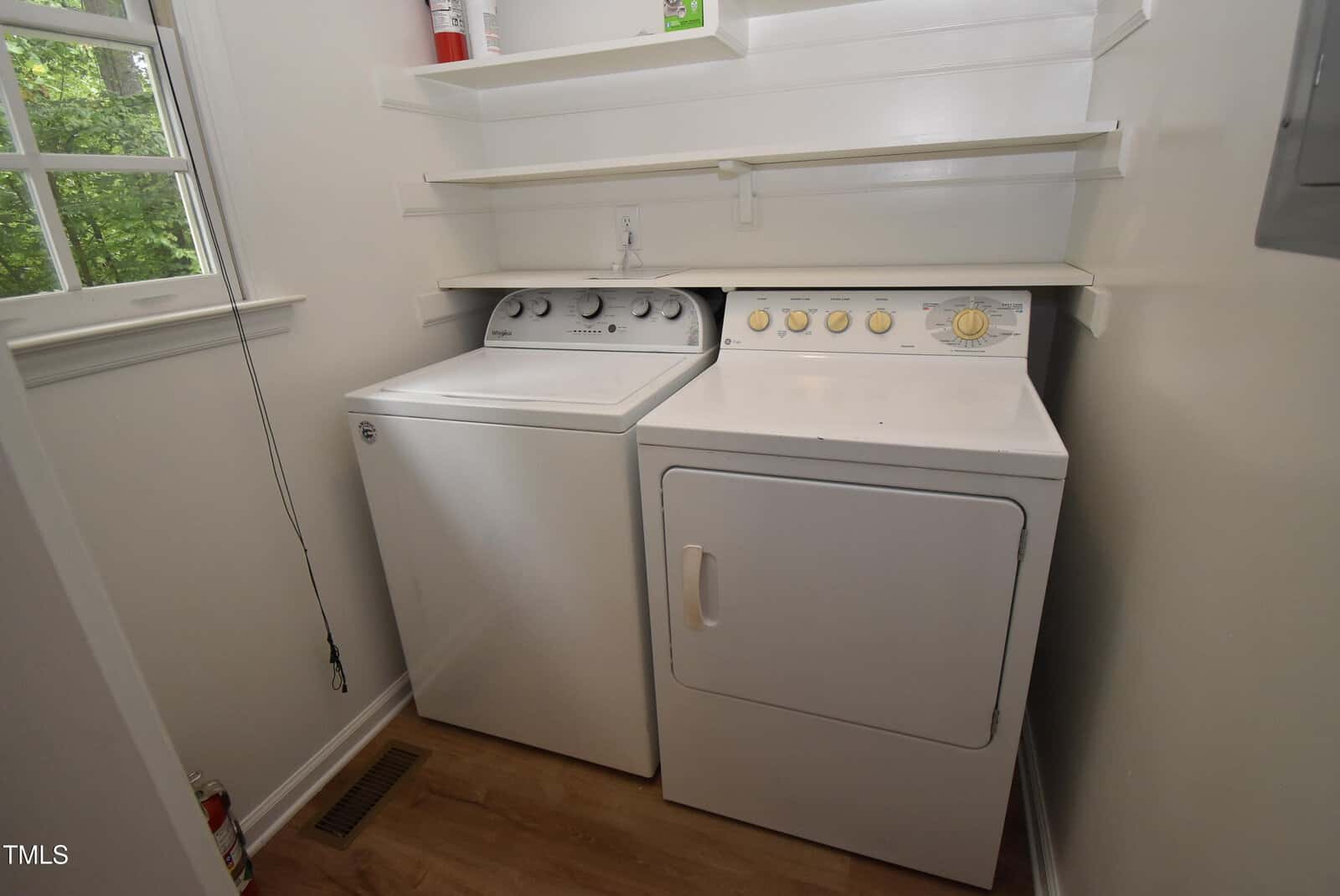 A laundry room with a white washing machine and dryer side by side, white shelves above them, a window to the left, and a wooden floor.