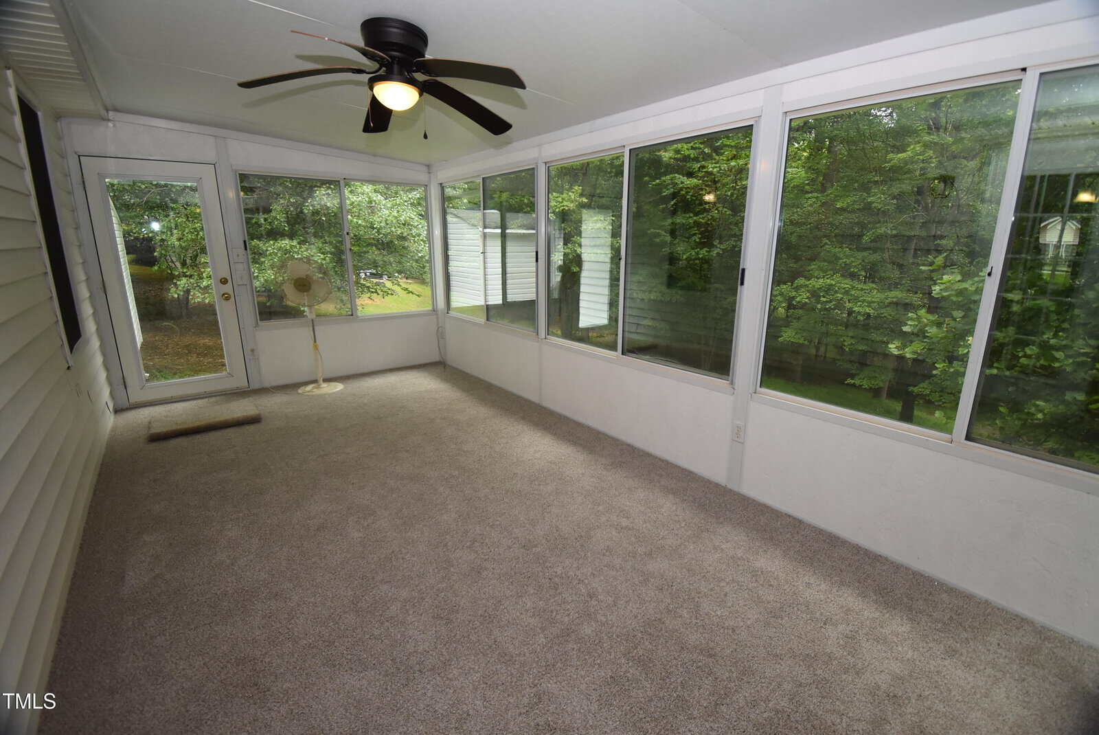Carpeted sunroom with large windows on two sides, a ceiling fan with light, glass door, and views of green trees outside. White walls and trim, with a floor vent and a small lamp in the corner.
