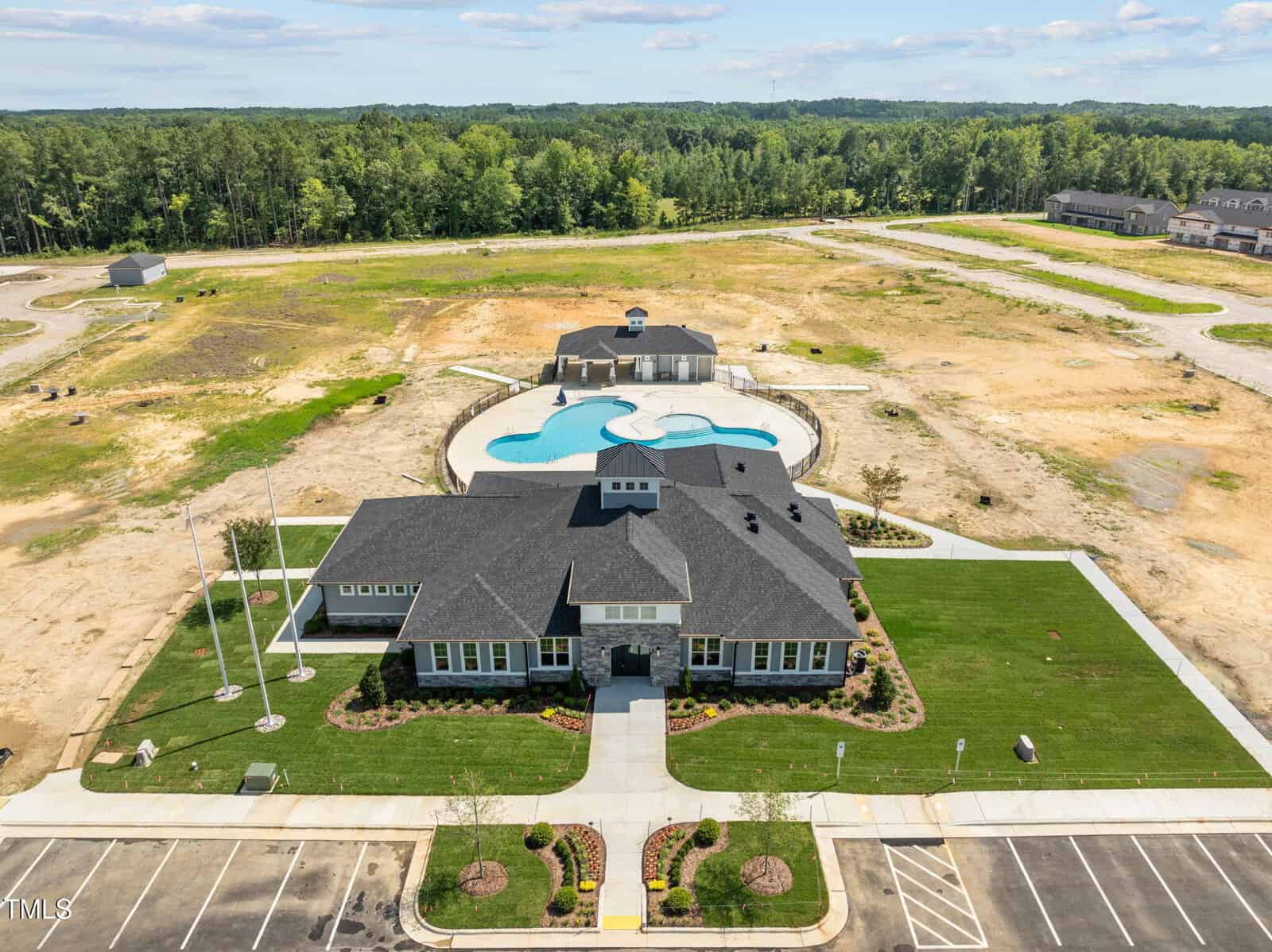 Aerial view of a modern community clubhouse with a swimming pool and a landscaped lawn, surrounded by a mostly undeveloped area with dirt and sparse vegetation, bordered by a wooded area in the background.