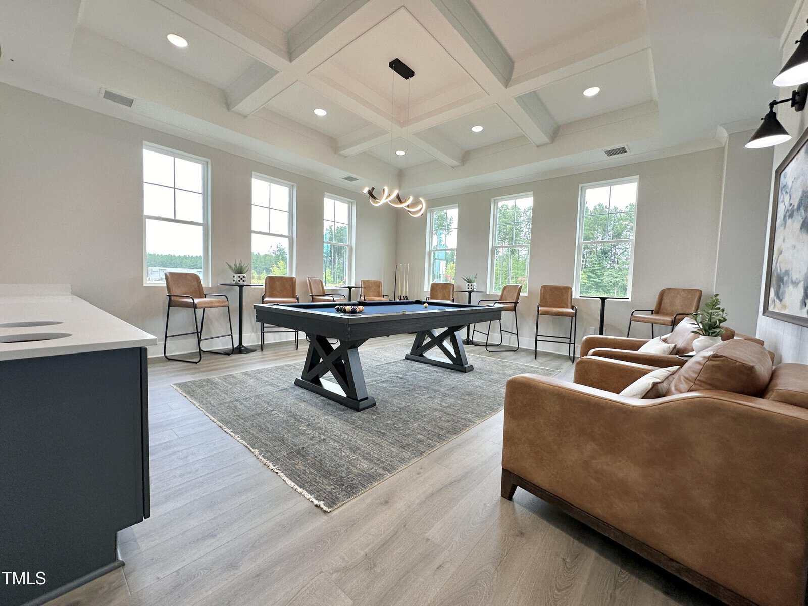 A modern game room with a pool table on a gray rug, tan leather chairs along the walls, a modern chandelier overhead, large windows letting in natural light, and light wood flooring.