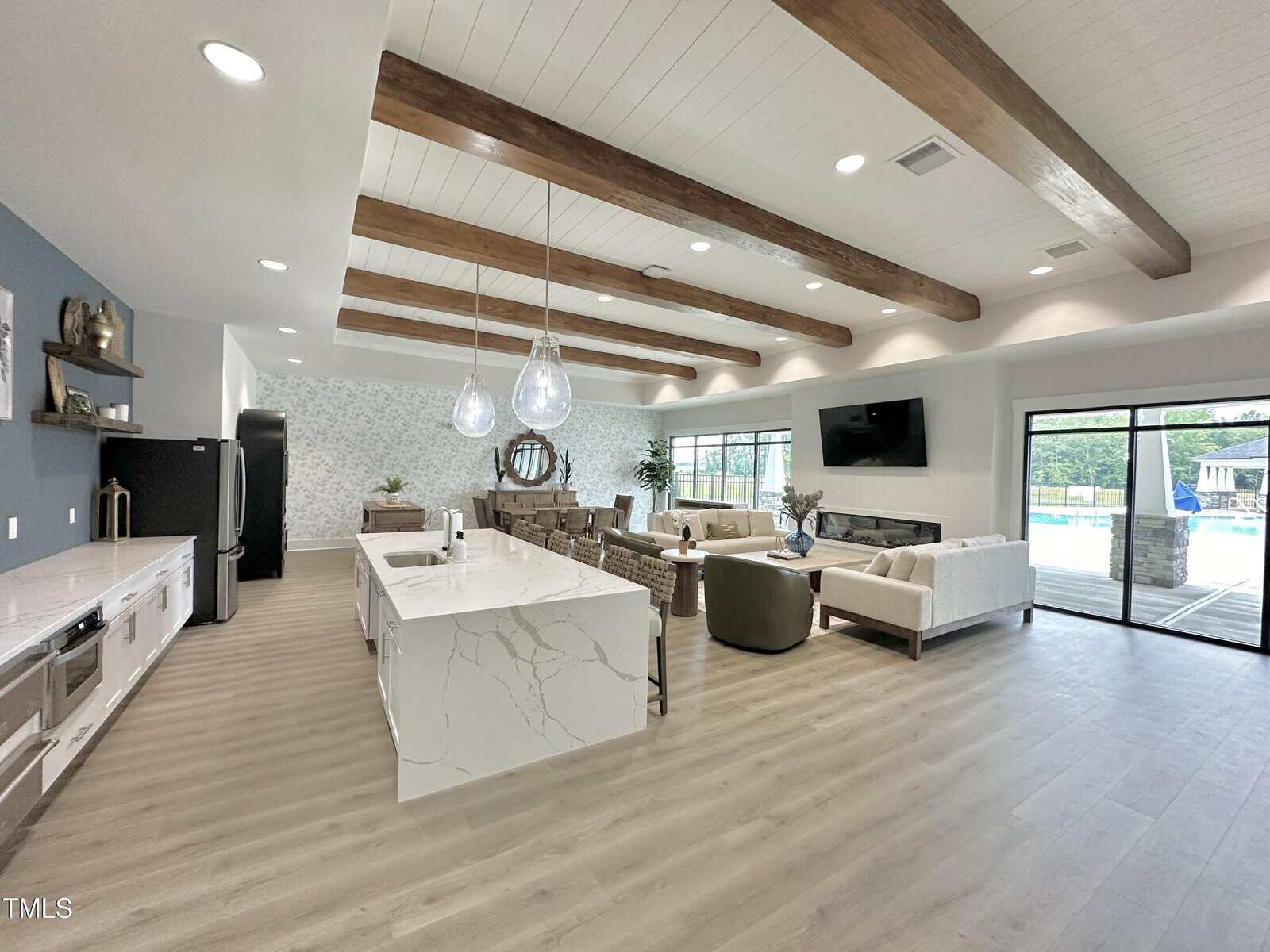 Spacious open-concept living area with wood beams on the ceiling, modern kitchen island, dining table, sofas, large wall-mounted TV, and glass doors overlooking a pool and outdoor area.