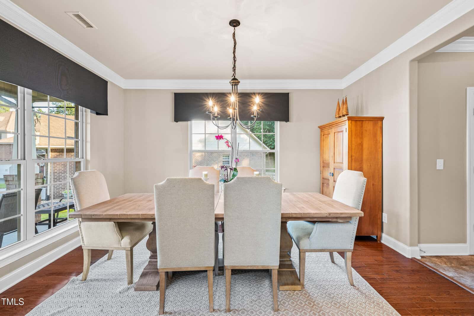 A dining room with a wooden table, six upholstered chairs, a chandelier above the table, large windows on one wall, light-colored walls, a wooden cabinet, and hardwood flooring with a rug under the table.