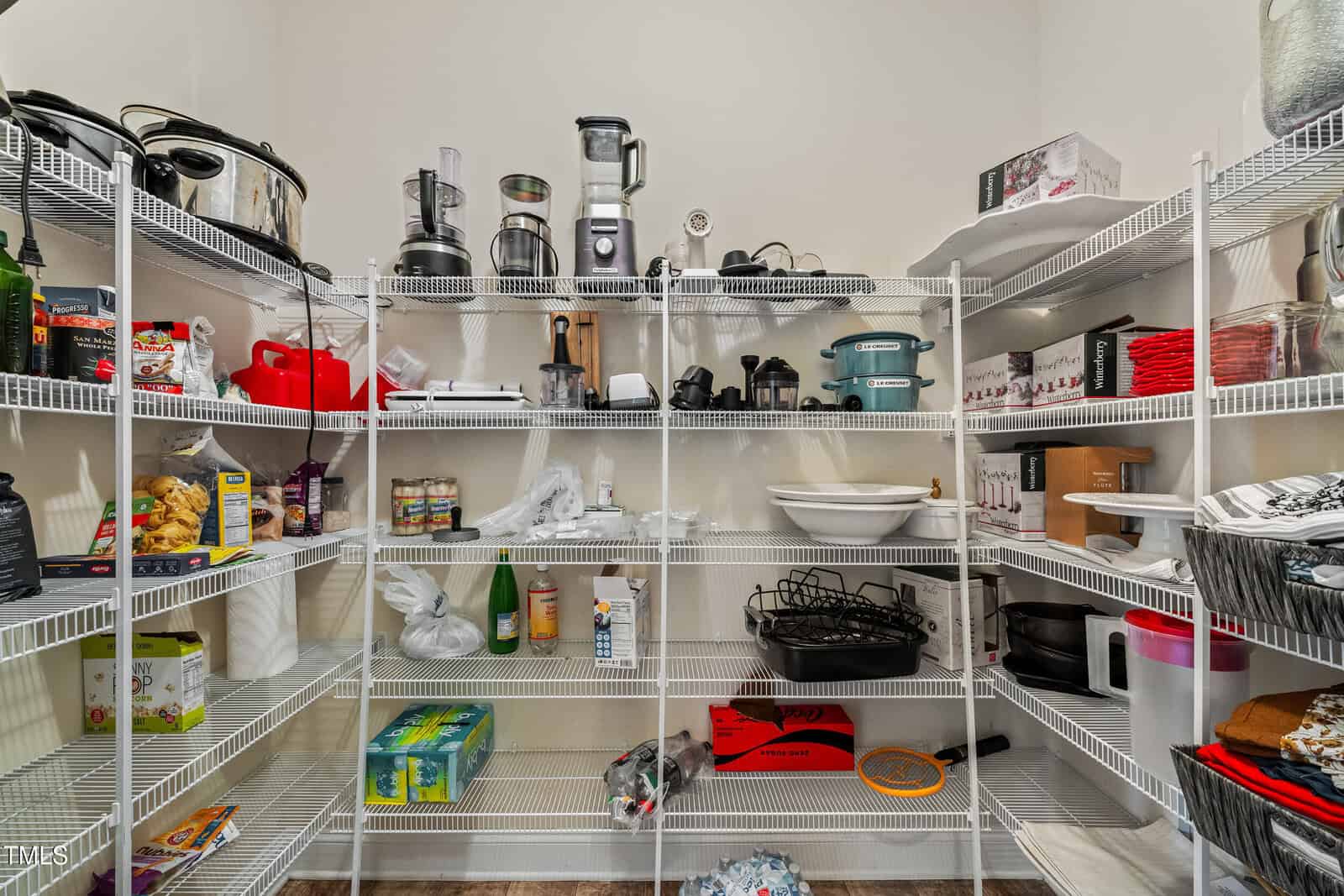 A pantry with white wire shelves holding kitchen appliances, food items, paper towels, cookware, and various household supplies, all neatly organized against white walls.