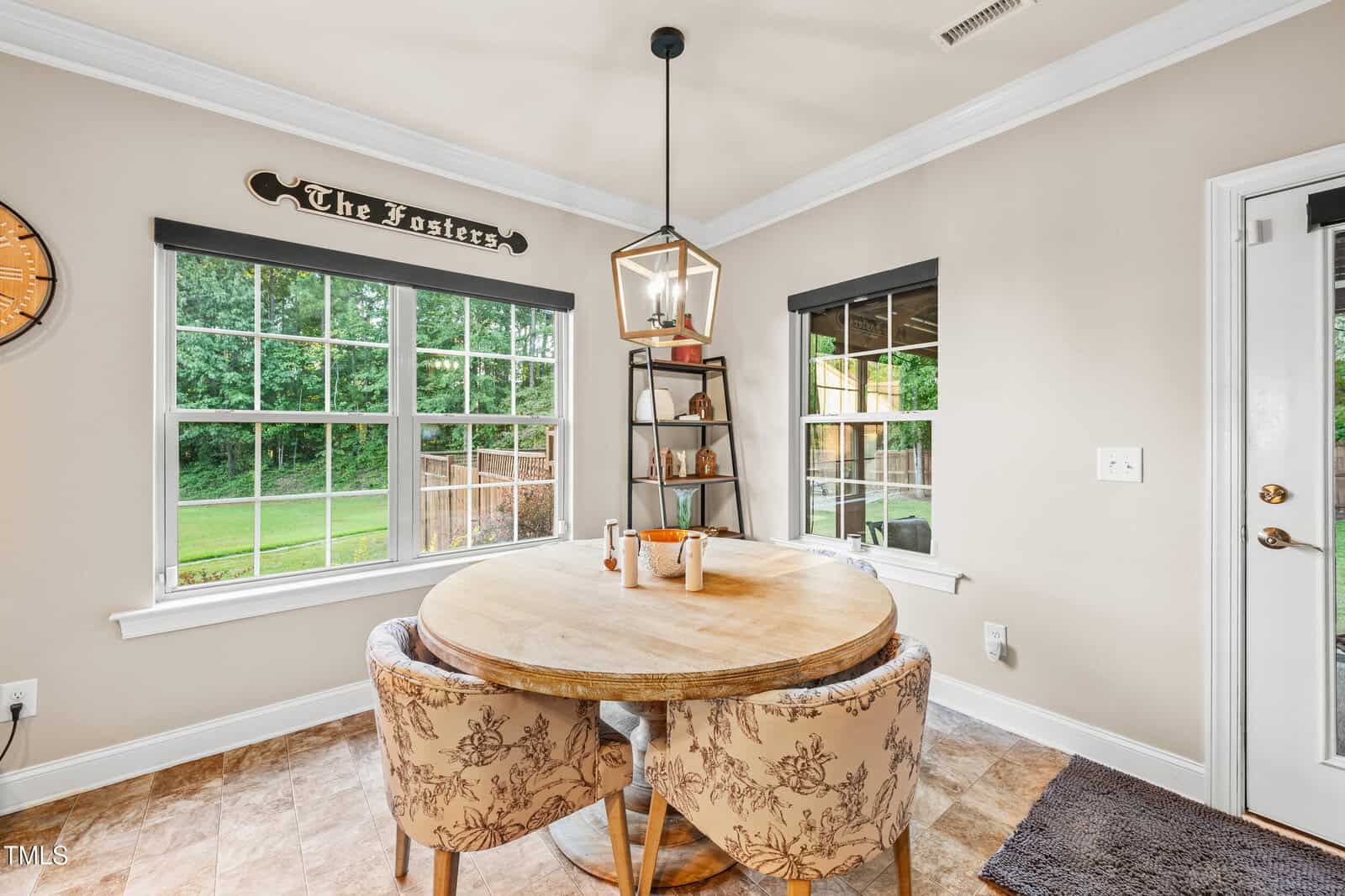 A cozy dining nook with a round wooden table, two upholstered chairs, a geometric pendant light, and large windows overlooking a green yard. A sign above the window reads “The Fosters.”.