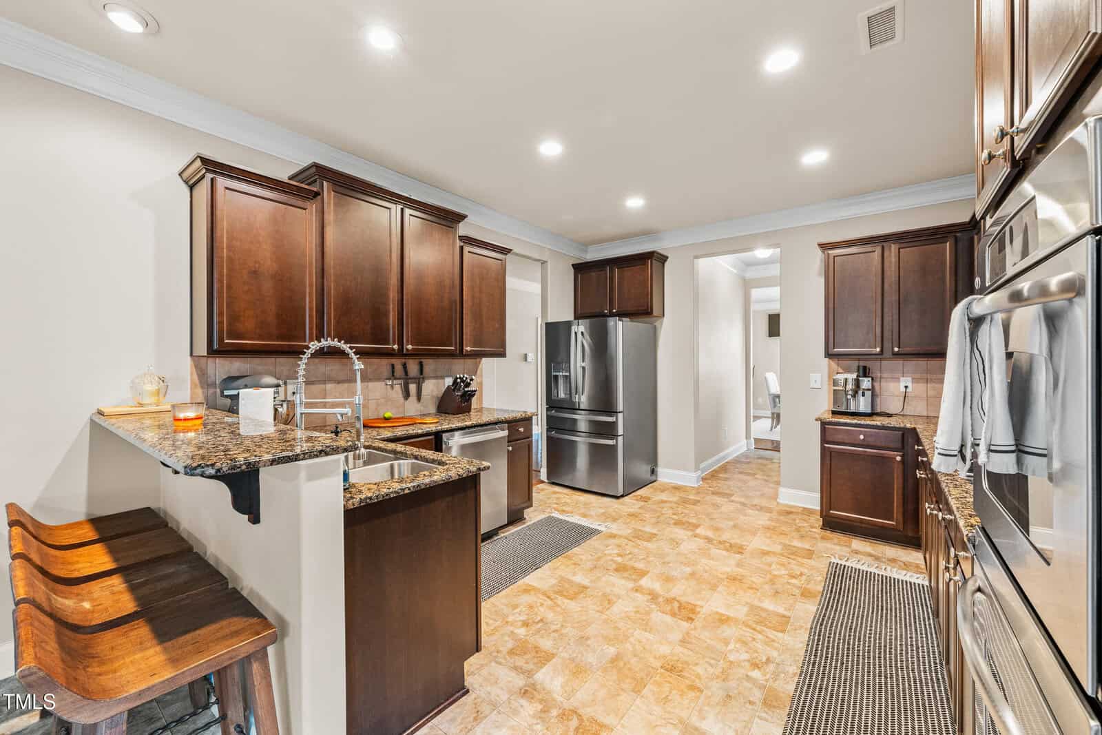 Modern kitchen with dark wood cabinets, stainless steel appliances, granite countertops, a double sink, two wooden barstools, and tile flooring. Bright overhead lighting and neutral walls create a clean, inviting space.