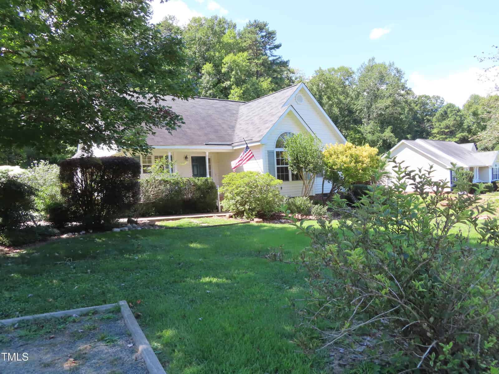 A single-story yellow house with white trim, an American flag on the porch, and a front yard with shrubs and trees under a clear sky. Another house is visible in the background to the right.