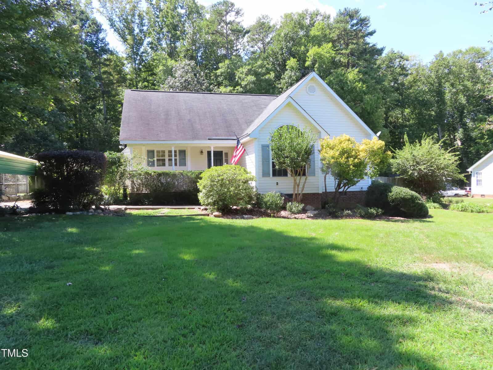 A white single-story house with a gable roof sits behind a well-kept lawn and various shrubs. An American flag hangs near the entrance. Trees fill the background under a clear, sunny sky.