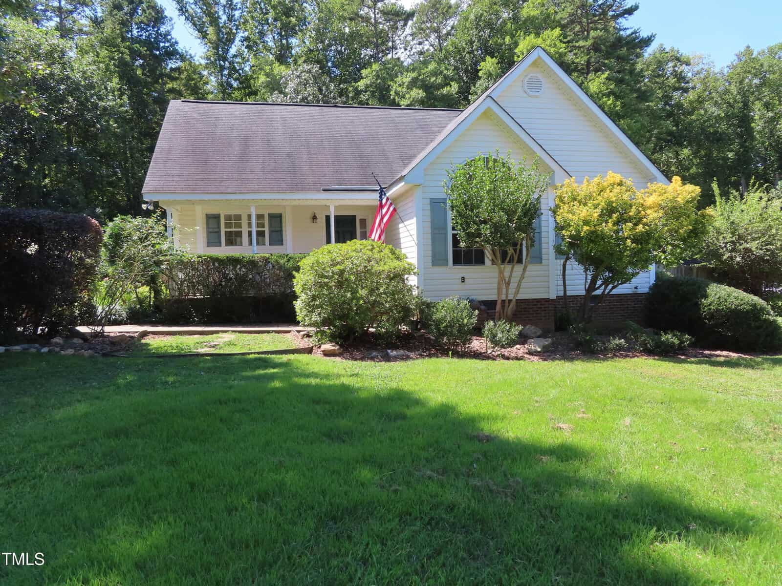 A single-story white house with a dark roof, front porch, American flag, and several bushes and trees in the yard. The lawn is green and well-maintained, with wooded areas in the background.