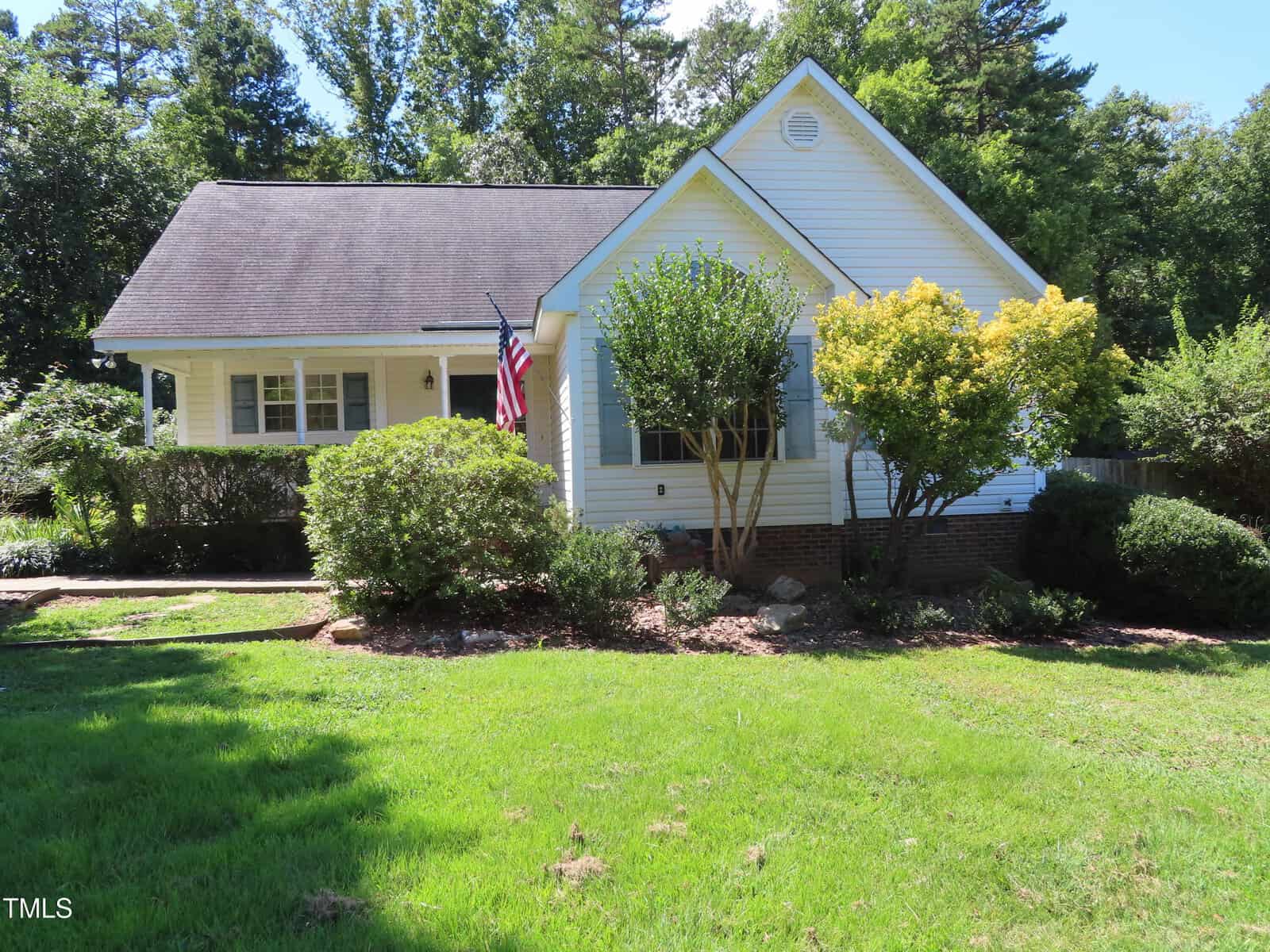 A single-story white house with a pitched roof on 5075 Range Road, Oxford, features a front porch, American flag, and various bushes and trees in the landscaped front yard under a clear blue sky.