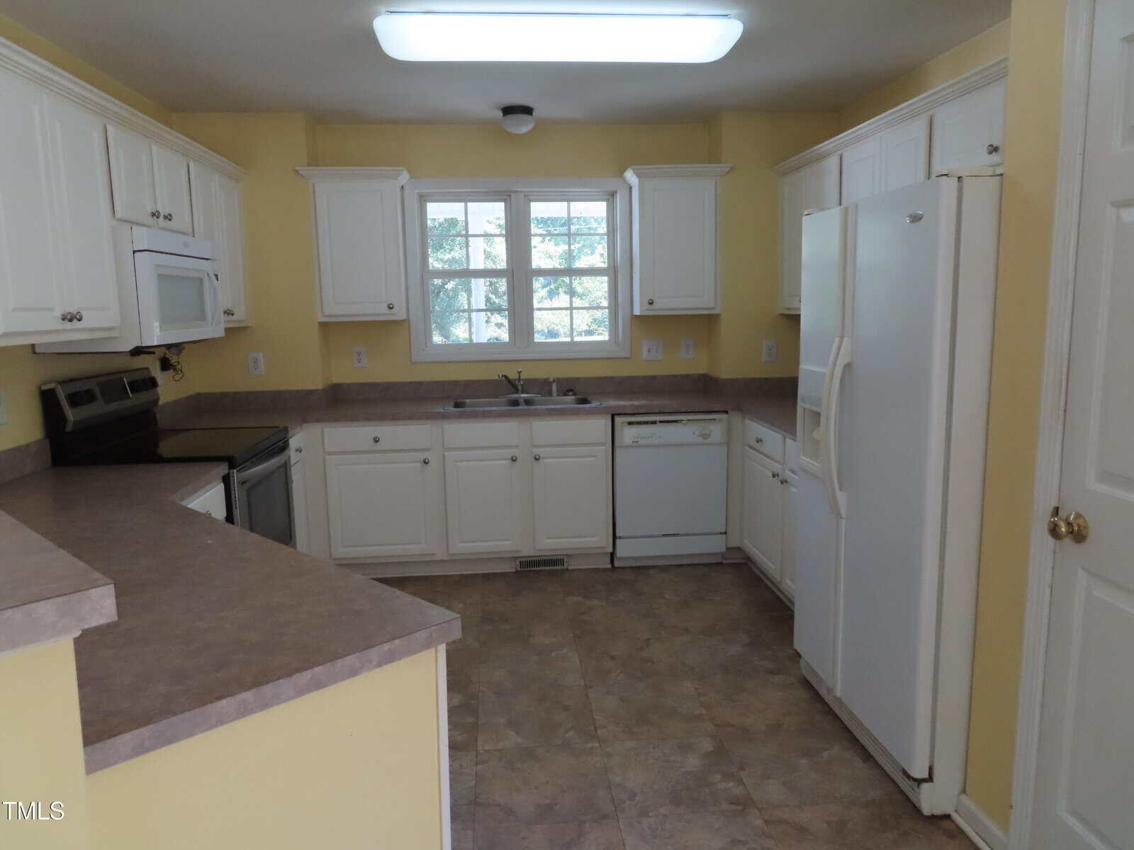 A kitchen with white cabinets, white appliances including a refrigerator, dishwasher, oven, and microwave, beige countertops, light brown tiled floor, yellow walls, and a window above the sink.