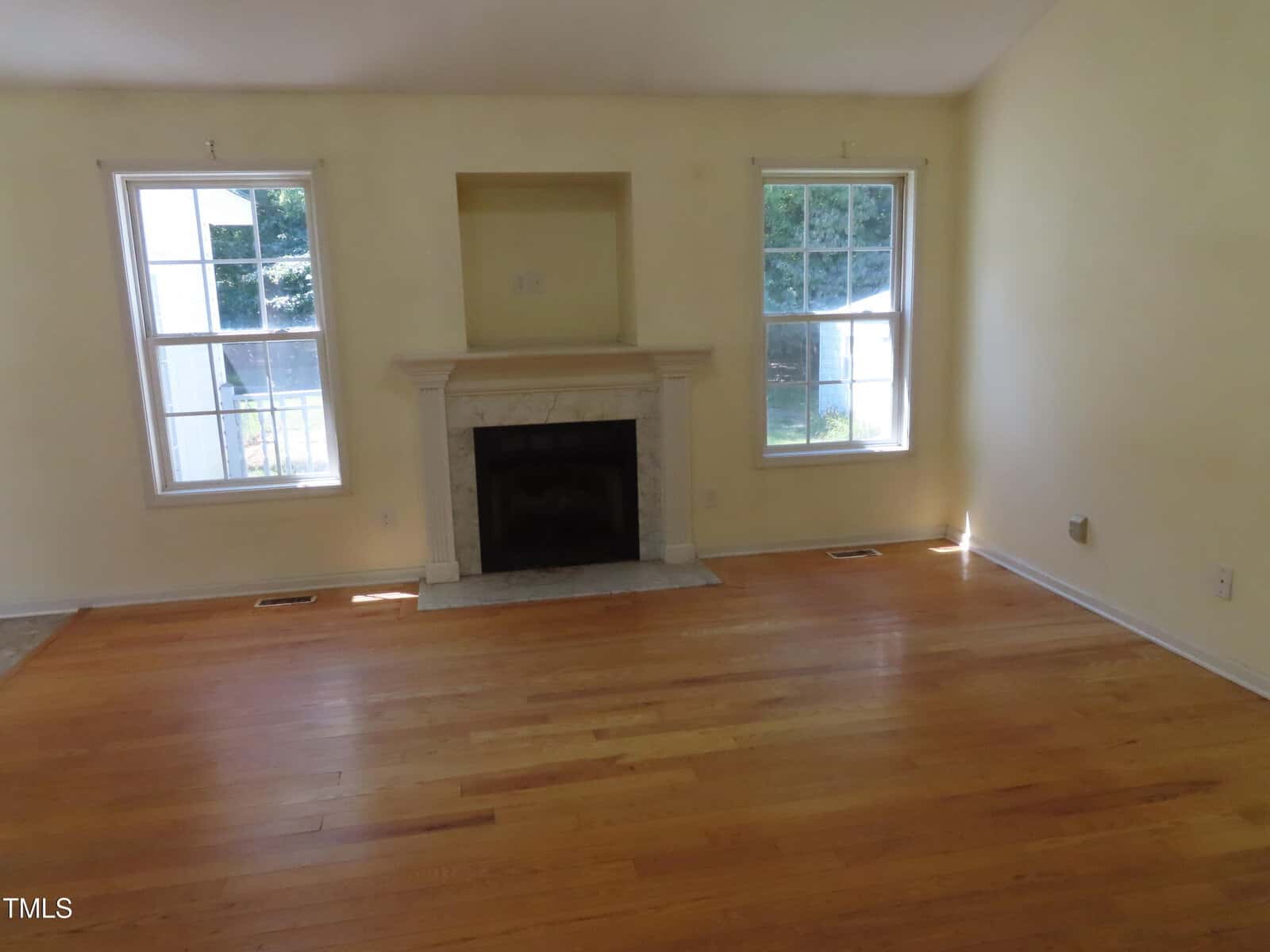 A room with light yellow walls, two tall windows, a white fireplace centered between them, and hardwood floors. Sunlight is coming through the windows, and the room is empty of furniture.