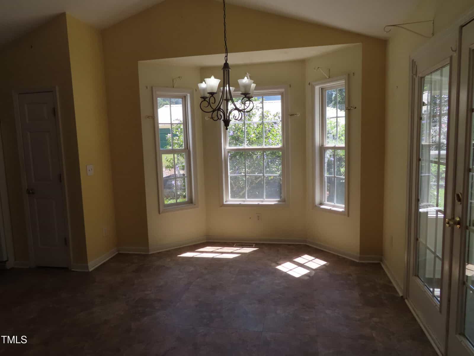 A bright, empty room with yellow walls, three large windows in a bay configuration, a glass-paned door, tile flooring, and a black chandelier hanging from the ceiling. Sunlight streams through the windows.