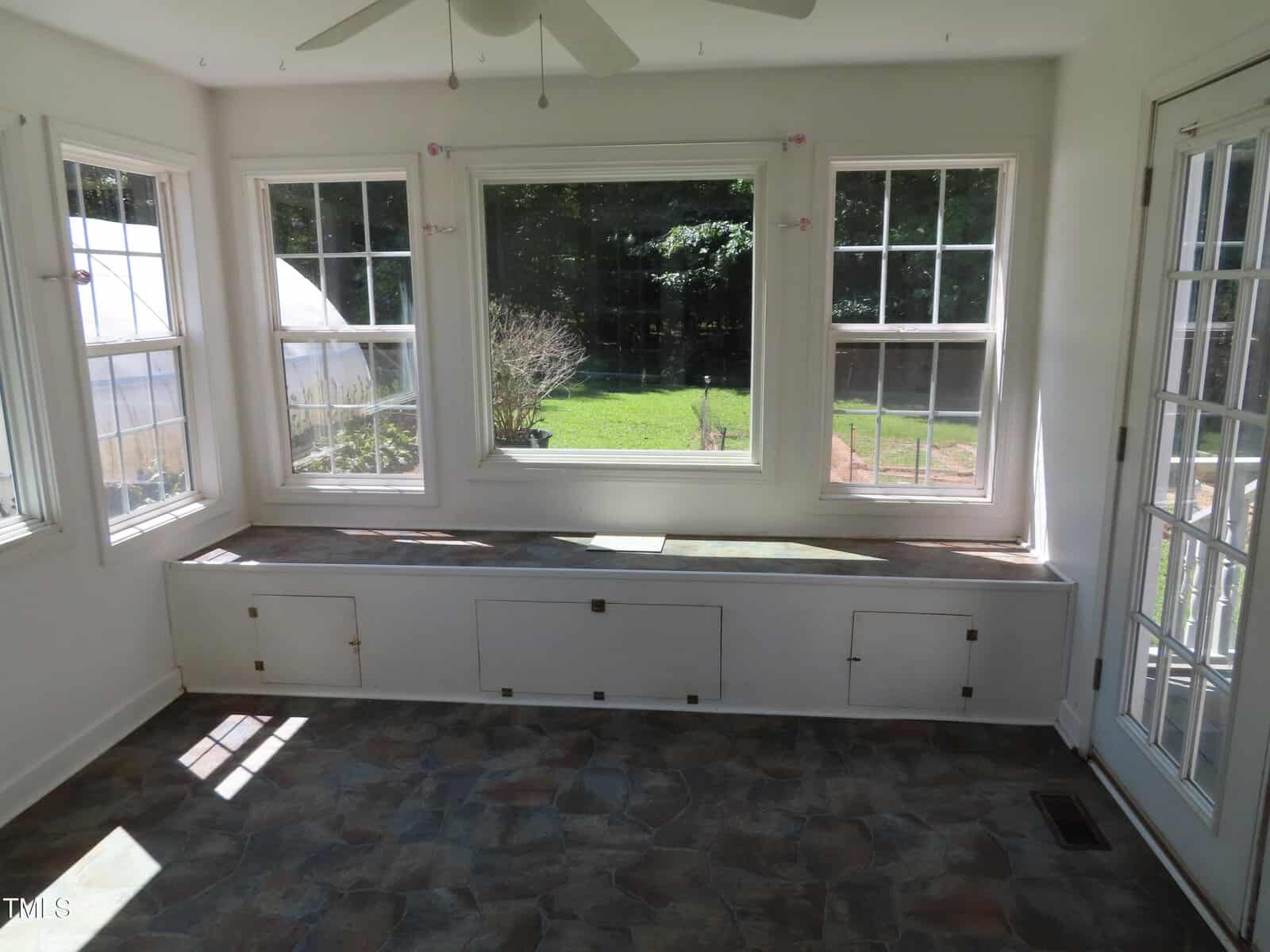 A sunroom with three large windows, a glass door, a ceiling fan, built-in bench storage along one wall, and stone-patterned tile flooring. The view outside shows a green lawn and trees.