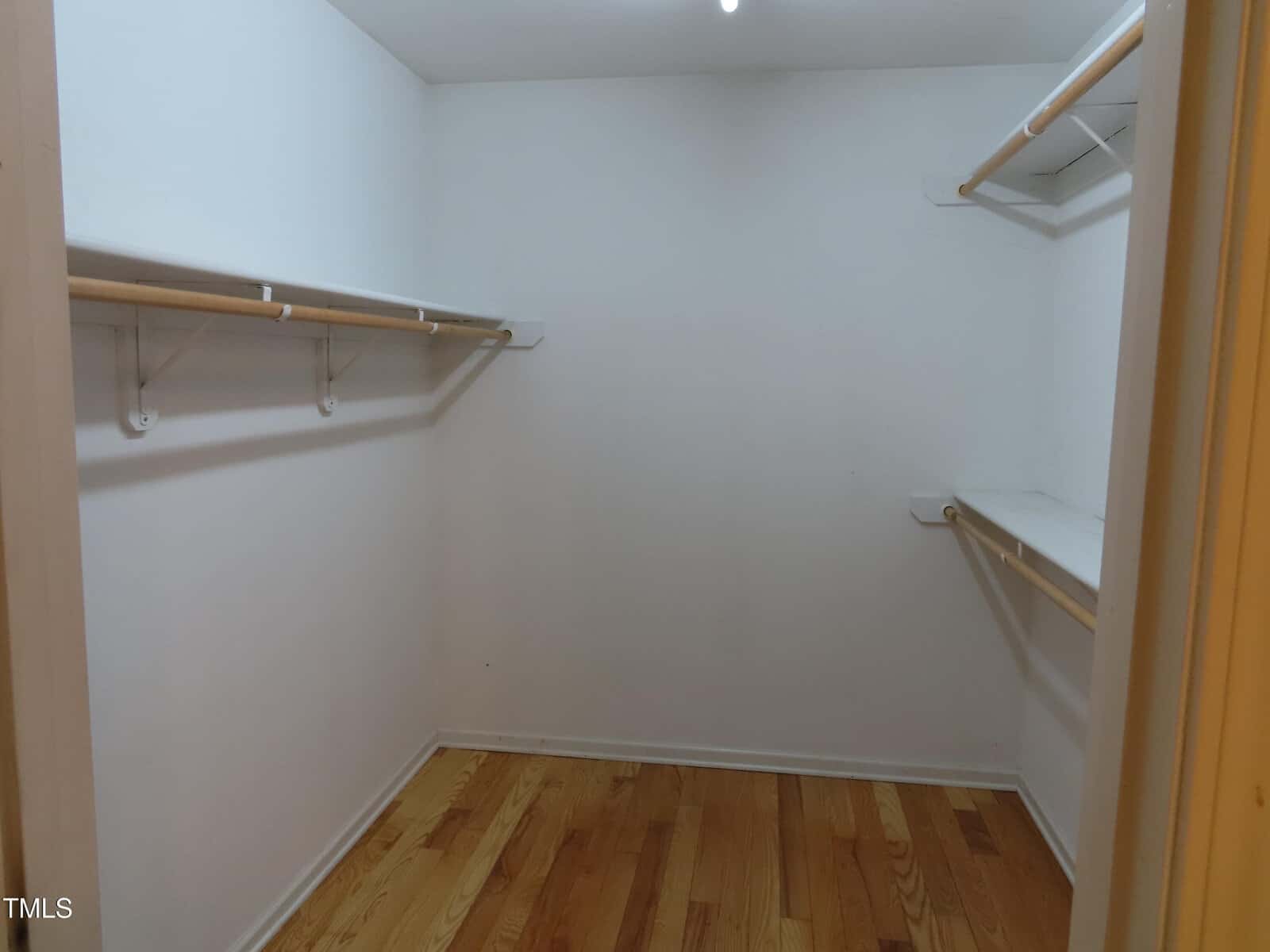 Empty walk-in closet with white walls, wooden floor, and multiple white hanging rods and shelves on both sides. Ceiling light is visible.