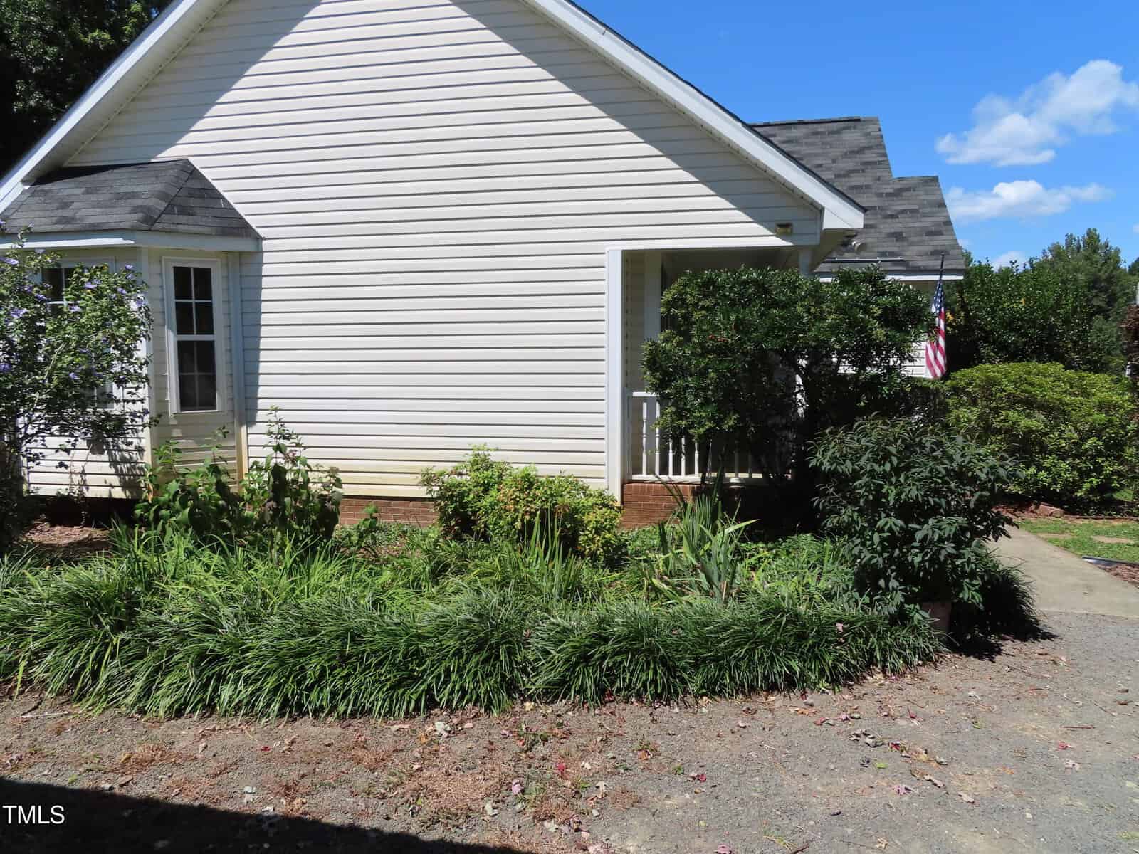 A white house with a small porch, green shrubs and plants along the front, a concrete walkway, and an American flag visible near the corner. The sky is clear and blue.