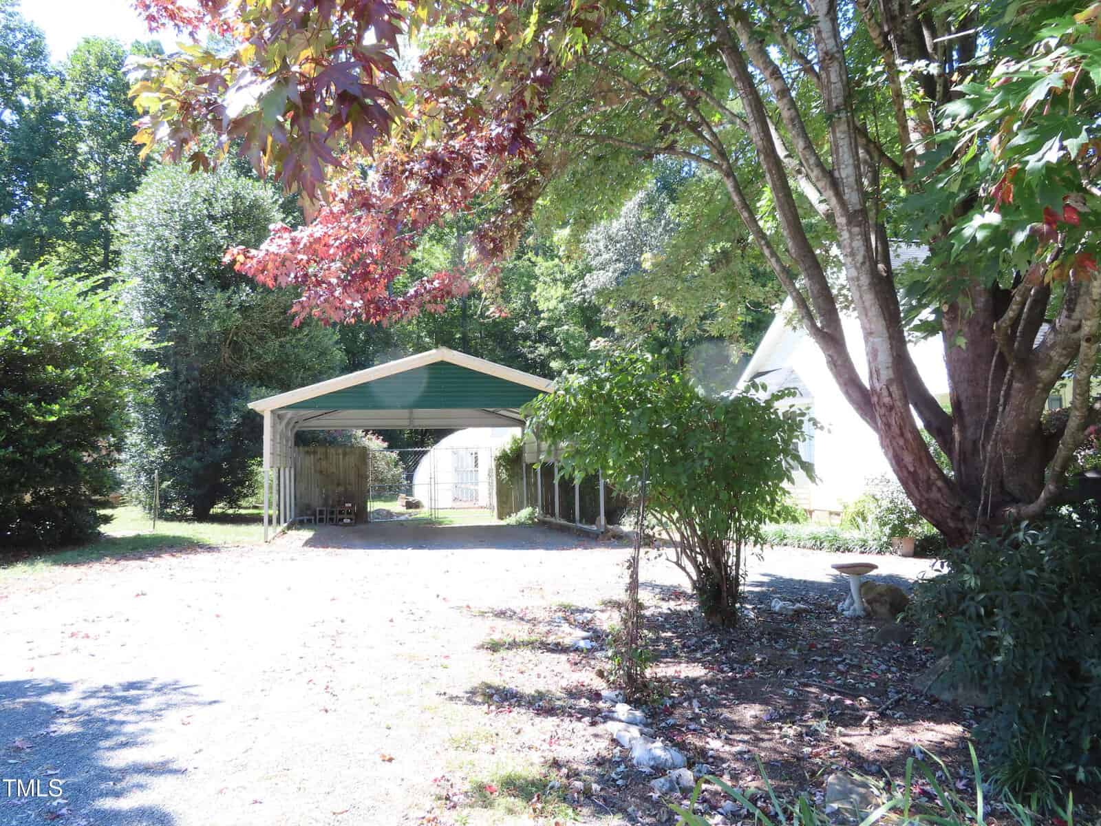 A green metal carport with a gravel driveway stands beside a white house. Large trees with green and reddish leaves provide shade, and sunlight casts dappled shadows on the ground.