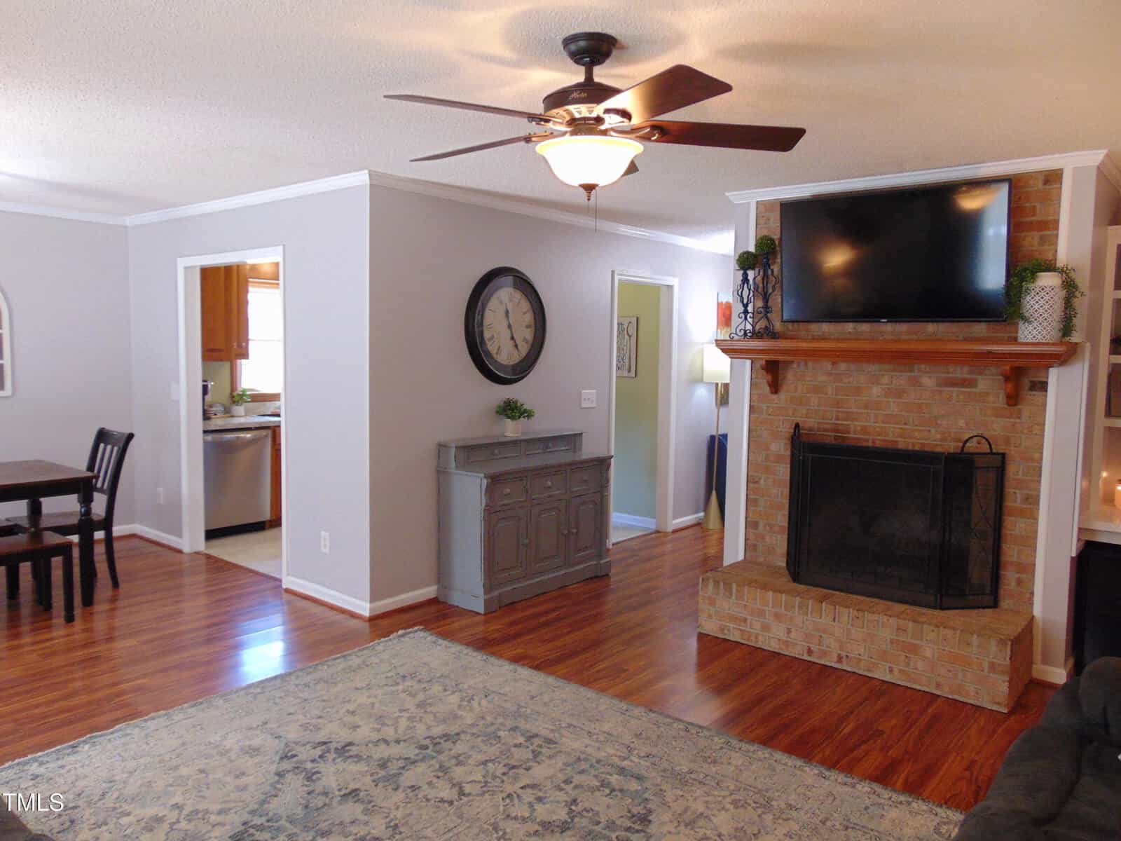 A cozy living room with wood floors, a brick fireplace with a mounted TV, a ceiling fan, gray walls, a large rug, and a dining area visible in the background. A wall clock and decorative items are displayed.